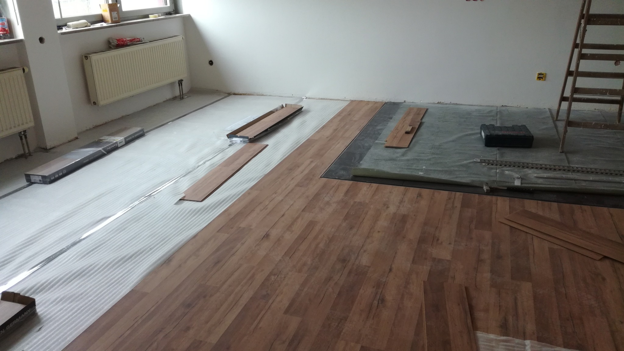 Podłoga w trakcie układania paneli, widoczne podłoże izolacyjne, narzędzia i częściowo ułożone panele w odcieniach brązu. W tle grzejnik i drabina.