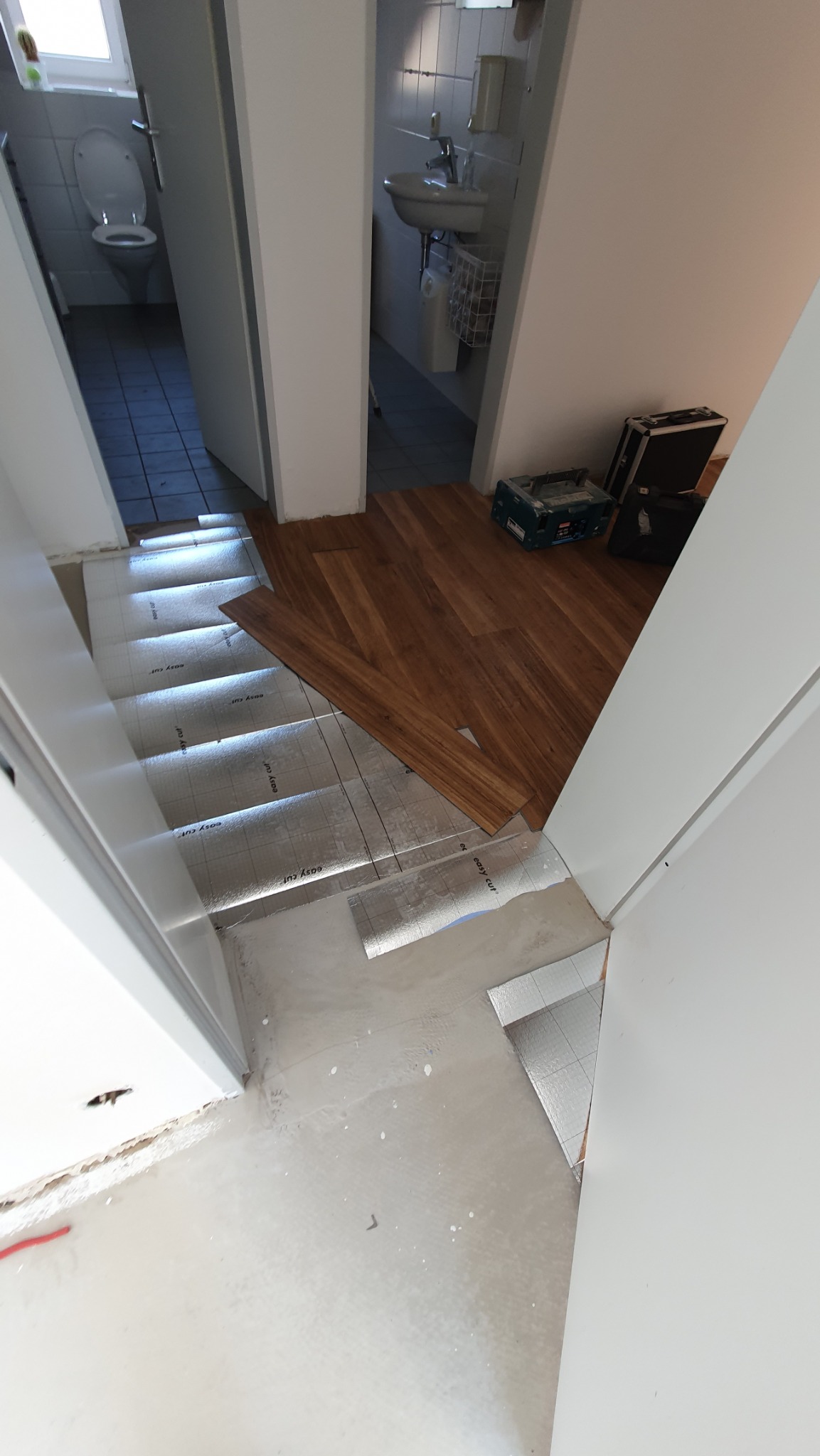 Fragment podłogi w trakcie układania paneli. Widoczne podłoże z folii, panele w odcieniach brązu i narzędzia. W tle widoczne pomieszczenia łazienek.