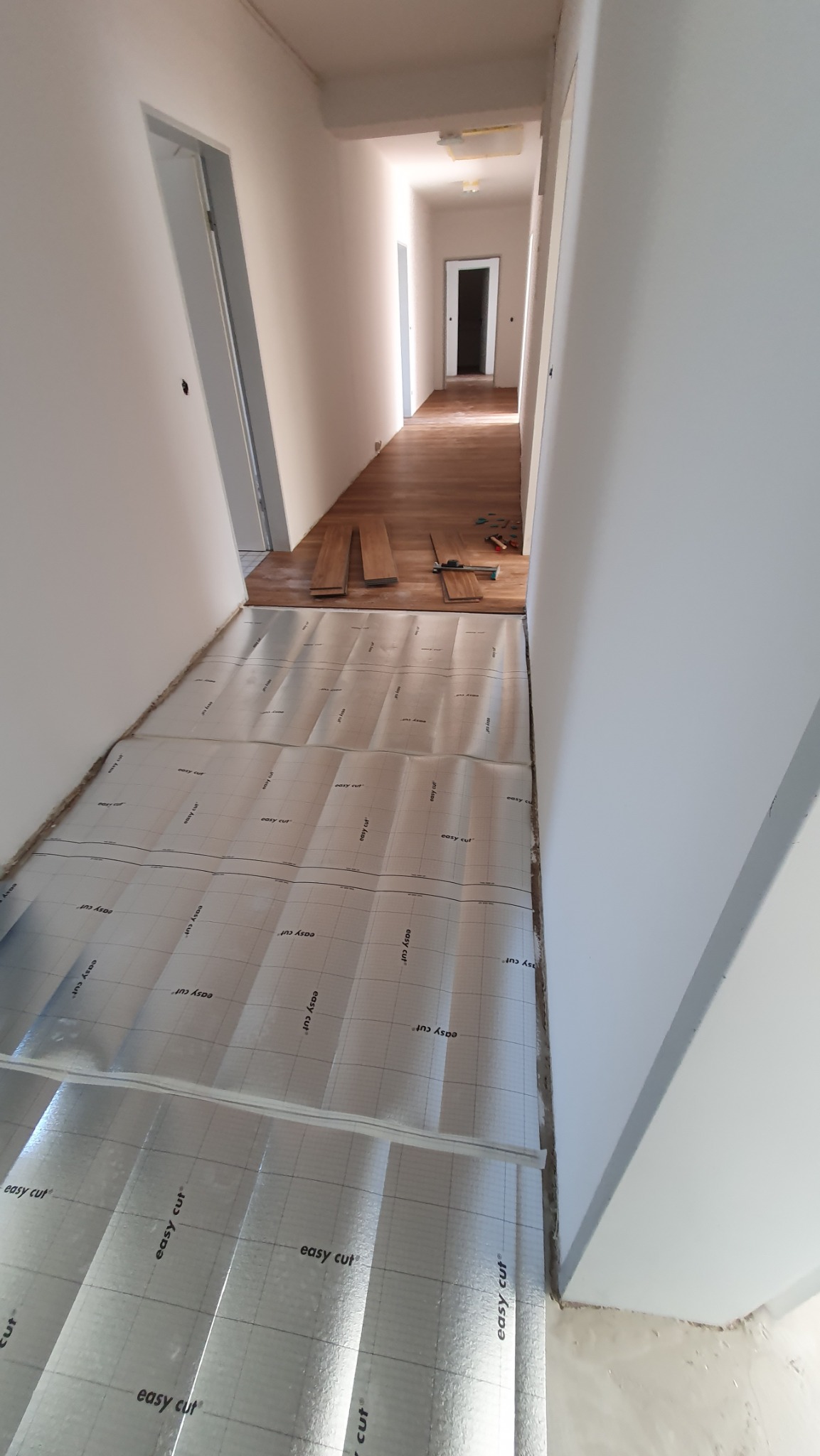 Korytarz w trakcie prac: podkład pod panele rozłożony, w tle ułożone panele podłogowe i narzędzia. Widok w perspektywie, jasne ściany.