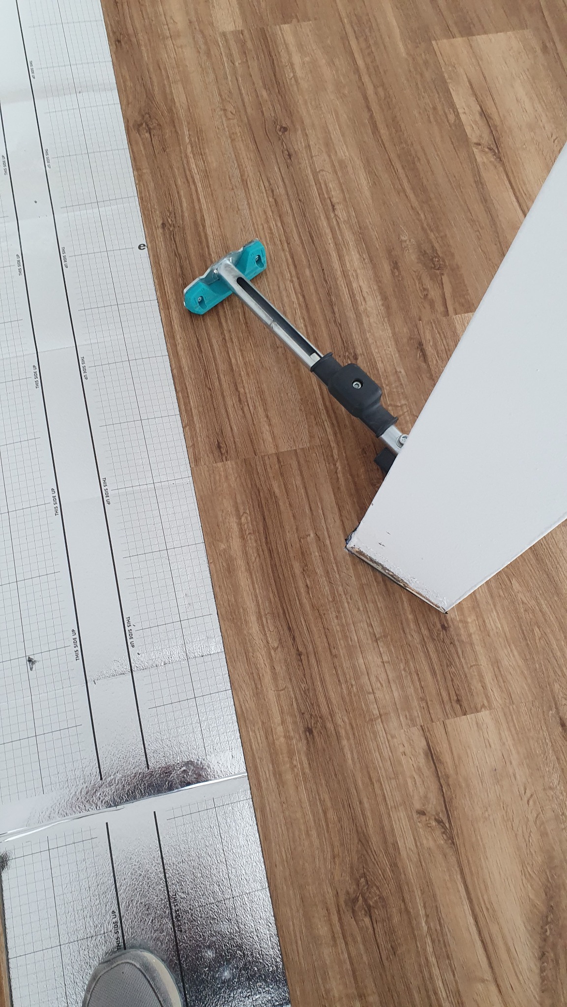 Przygotowanie podłoża: folia izolacyjna z nadrukiem siatki, narzędzie do dociskania paneli i fragment panela, podłoga imitująca drewno. Widok z góry.