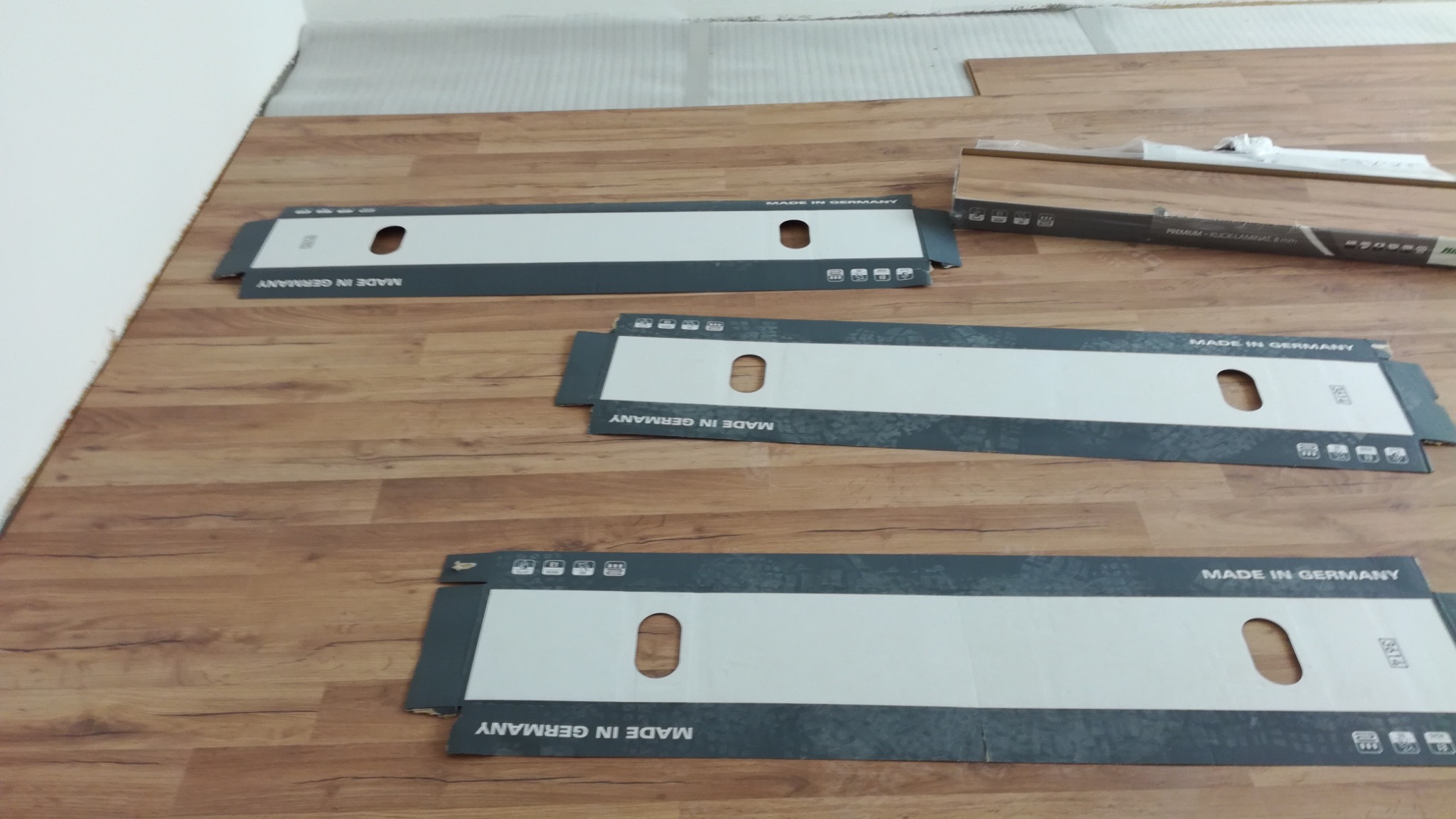 Układanie paneli: podłoga z paneli o wzorze drewna, widoczne kartony po panelach z napisem 'Made in Germany', fragment ściany w tle.