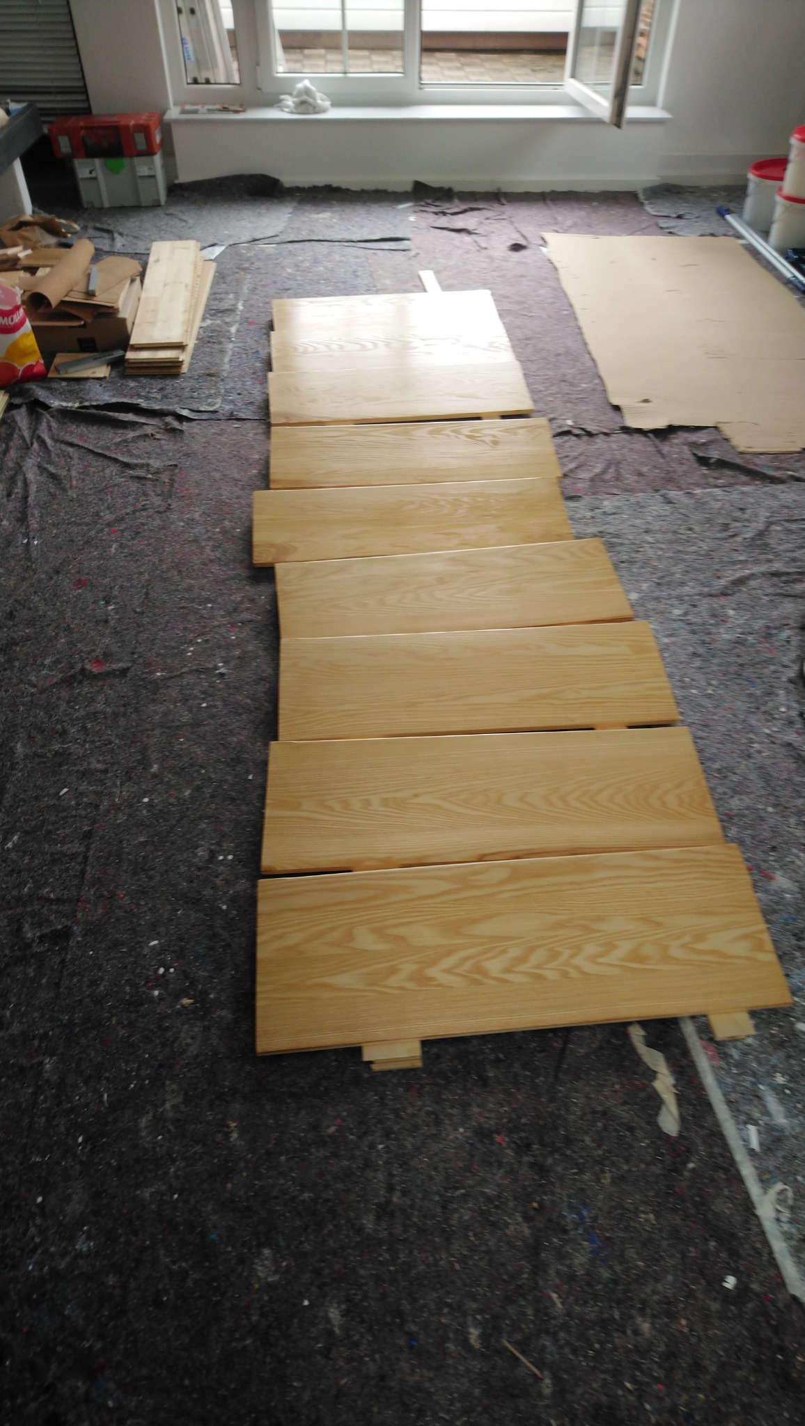 Jasne panele ułożone rzędami na podłodze zabezpieczonej szarą folią malarską, przygotowane do montażu. Widoczne okno w tle i materiały budowlane.