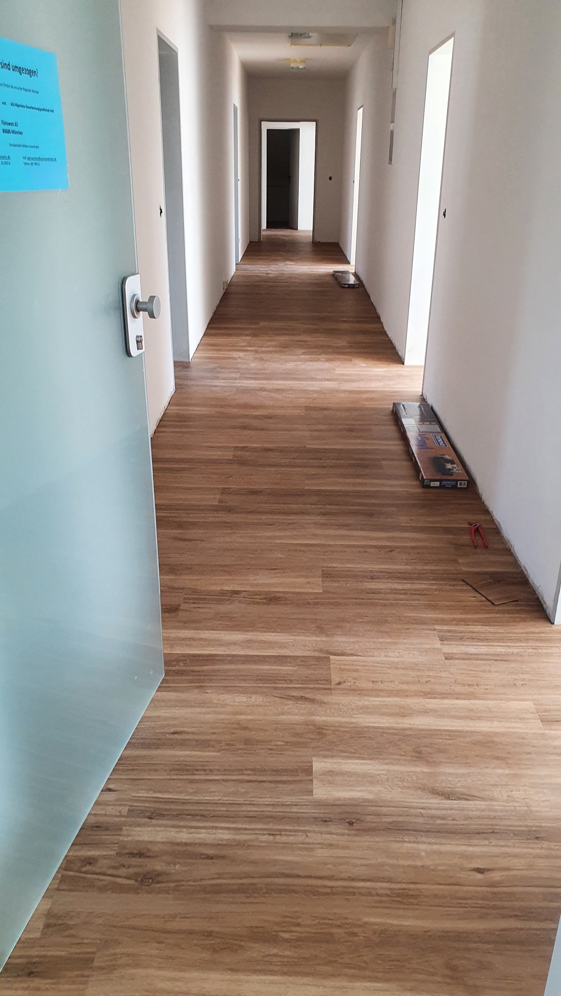 Długi korytarz z nową, drewnopodobną podłogą z paneli. Widoczny fragment paczki paneli i narzędzia. Otwarta szklana drzwi po lewej stronie kadru.