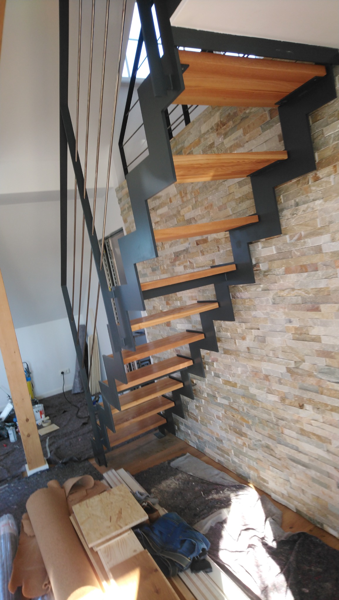 Nowoczesne schody z drewnianymi stopniami i metalową konstrukcją, zamontowane przy ścianie wyłożonej dekoracyjnym kamieniem. Widoczne materiały budowlane w tle.