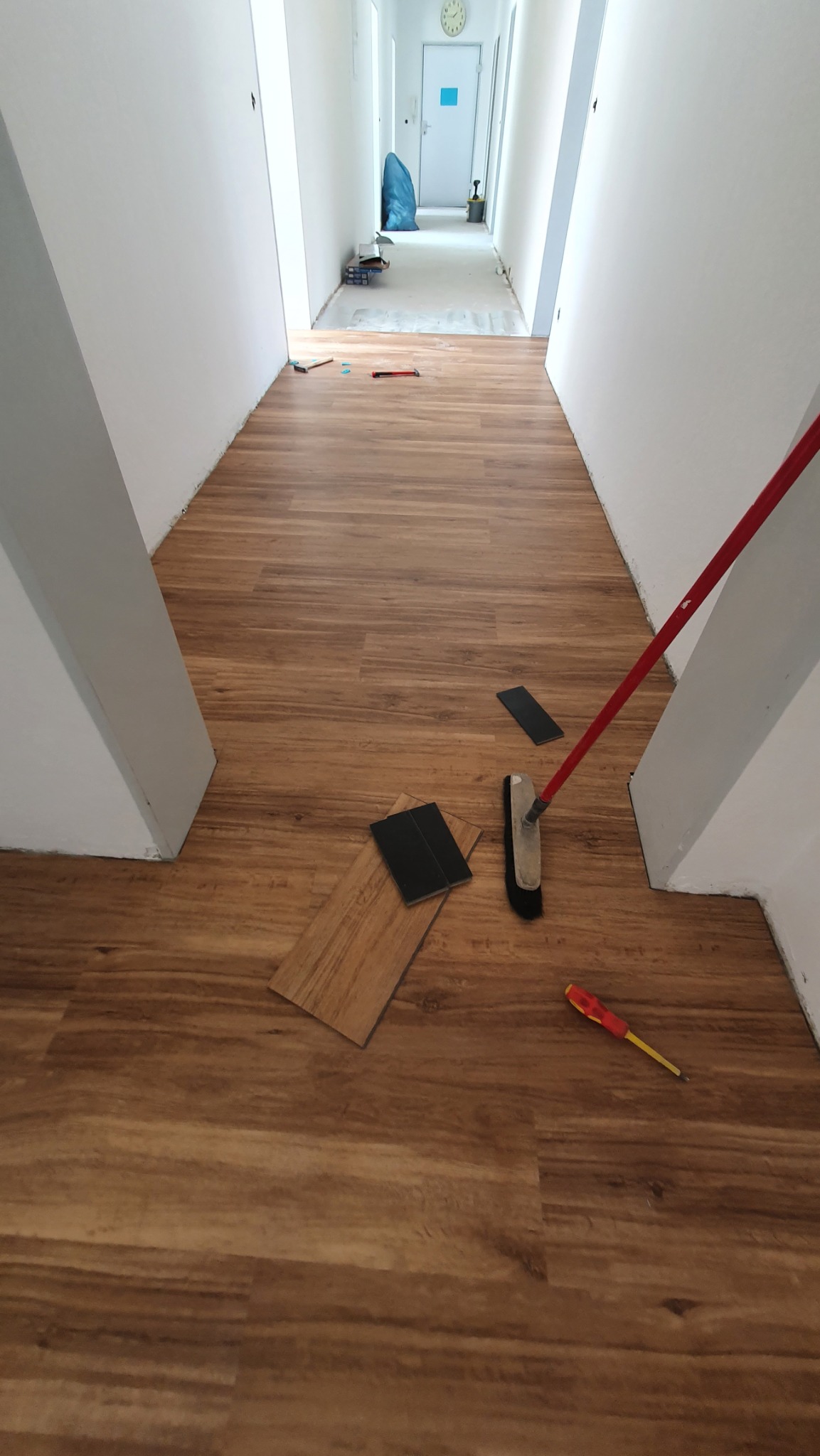 Korytarz w trakcie układania paneli podłogowych. Widoczne panele o wzorze drewna, narzędzia pracy: miotła i śrubokręt. Część podłogi jeszcze nieukończona.