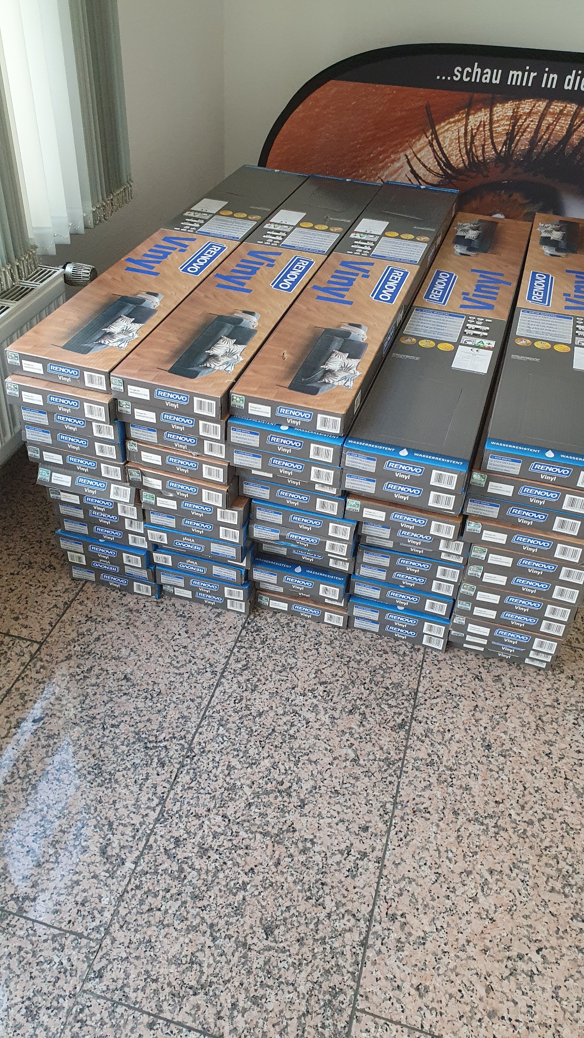 Stos opakowań paneli winylowych Renovo, ułożonych na podłodze z płytek. Widoczne okno z żaluzjami i fragment reklamy z okiem w tle.