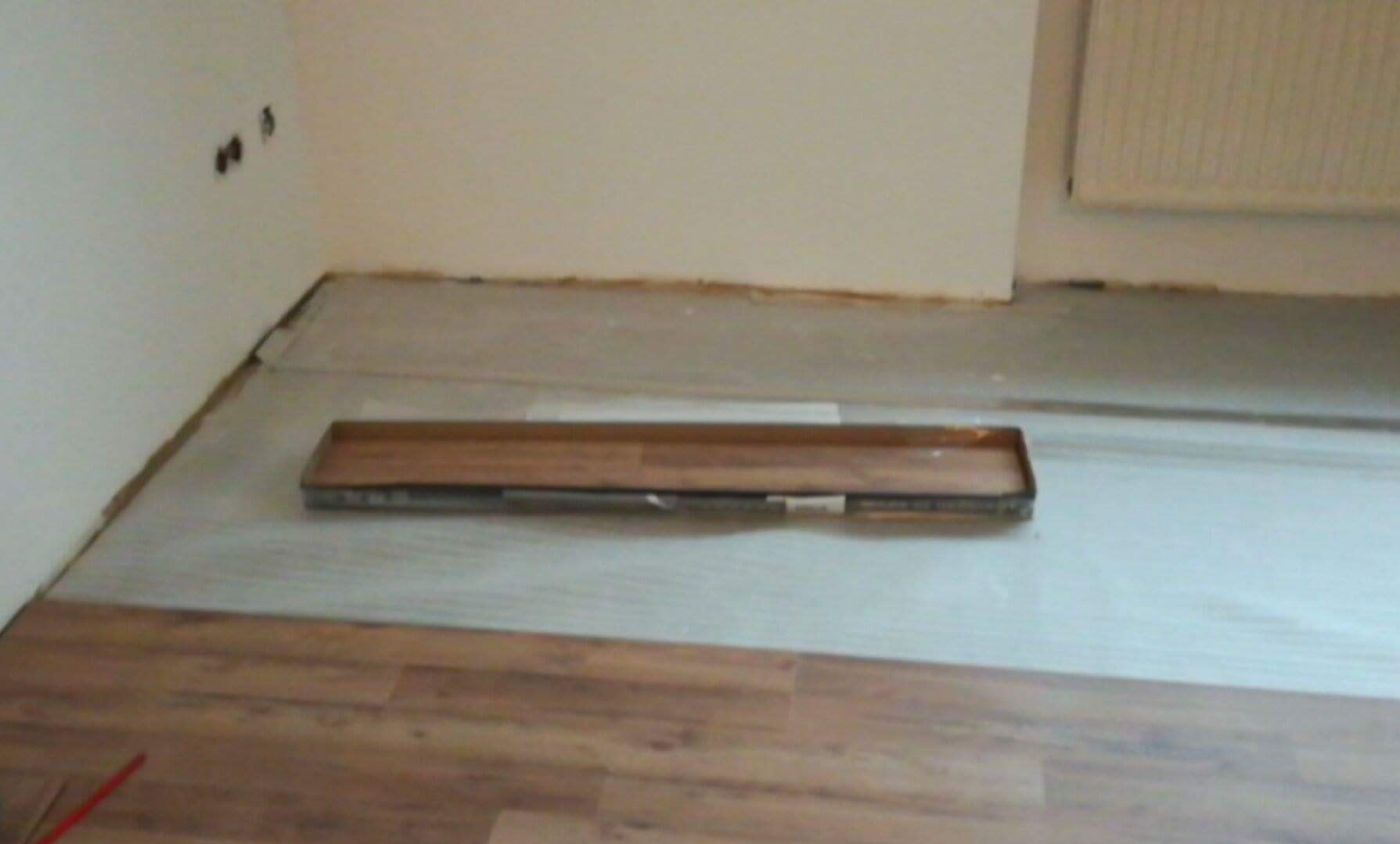 Fragment podłogi z ułożonymi panelami, widoczna paka paneli na podkładzie, ściana w tle z gniazdkami. Układanie w trakcie prac.