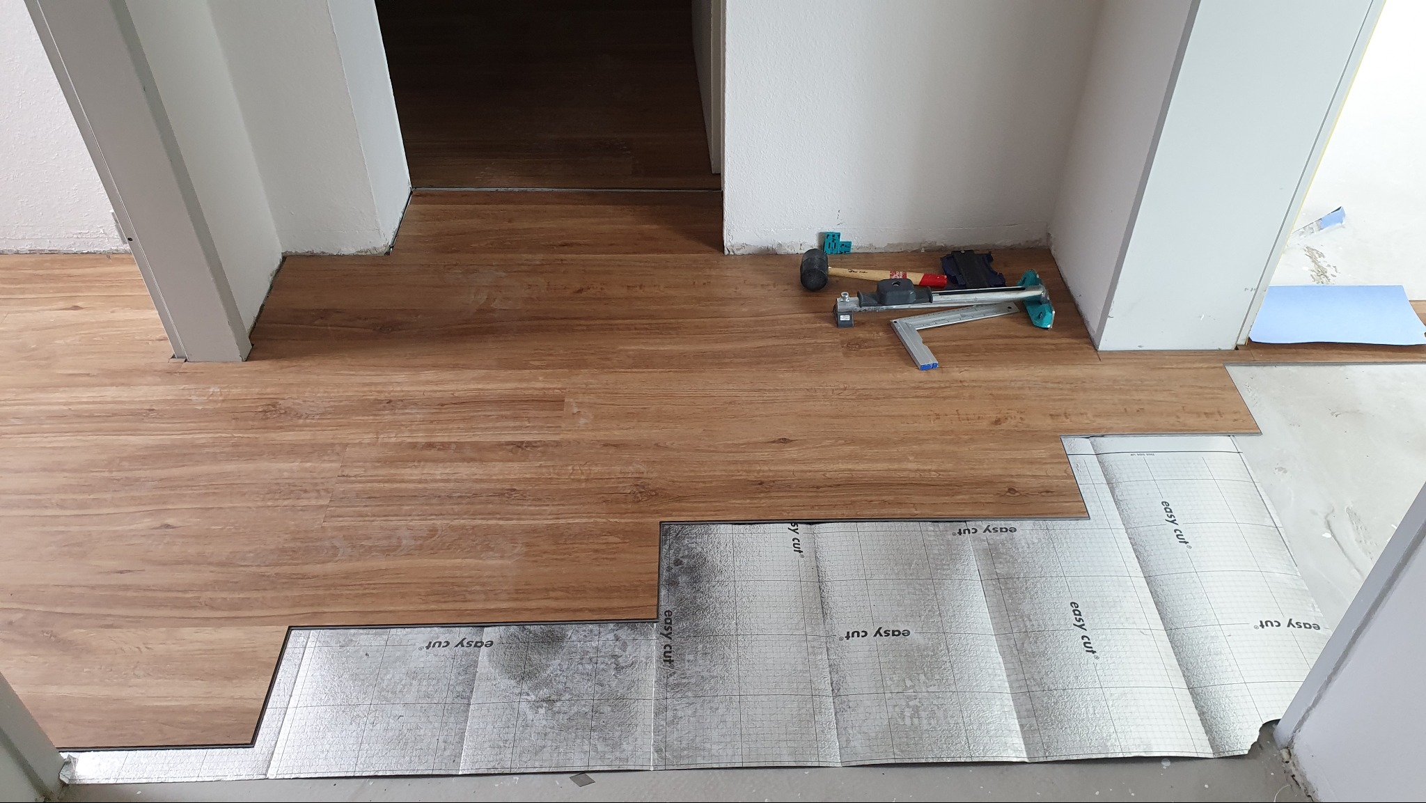 Proces układania paneli podłogowych z widocznymi narzędziami i podkładem izolacyjnym 'easy cut' w jasnym wnętrzu. Częściowo ułożona podłoga z paneli imitujących drewno.