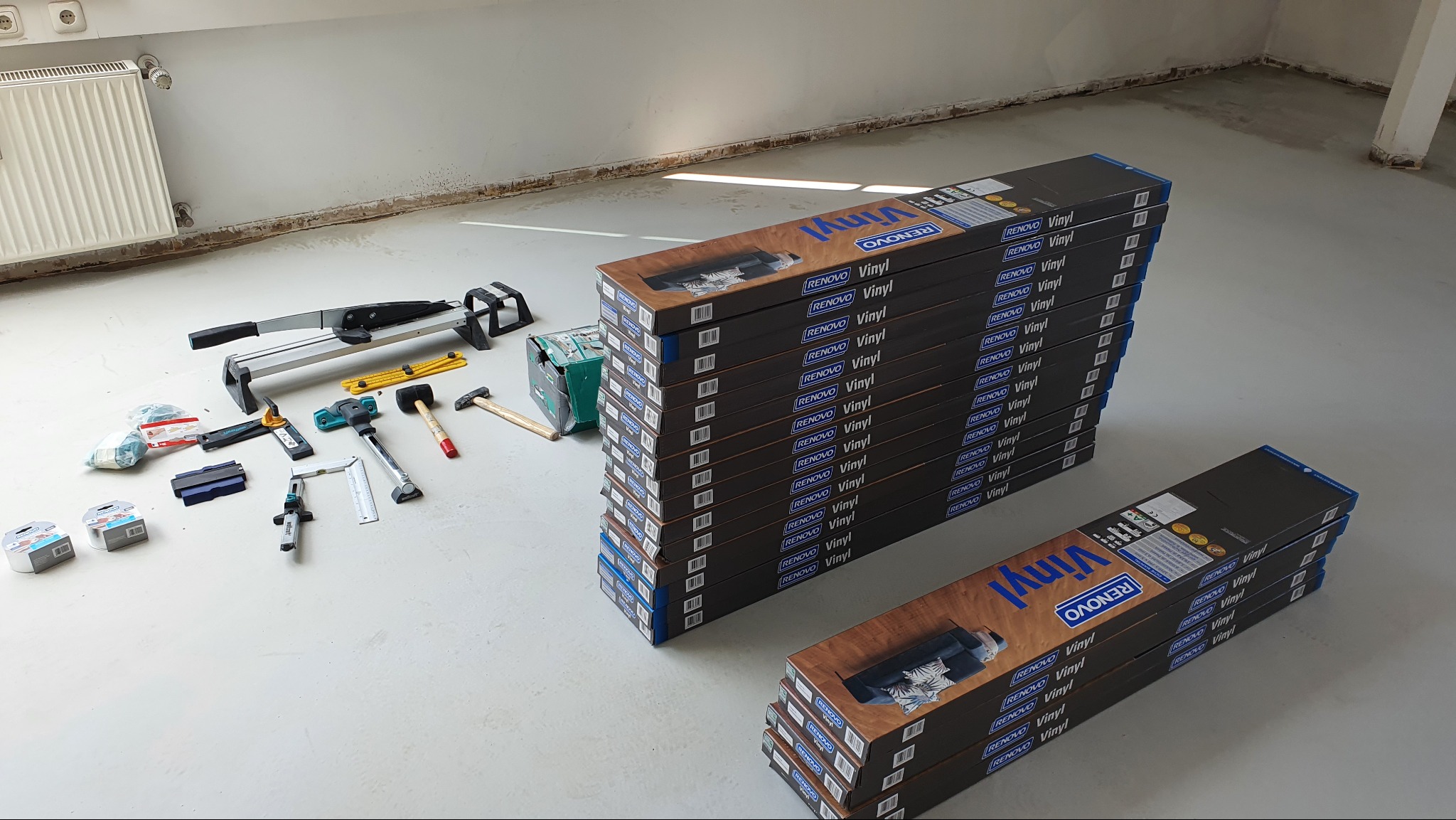 Przygotowanie do montażu paneli winylowych: narzędzia, akcesoria i opakowania paneli Renovo Vinyl ułożone na jasnej posadzce w pustym pomieszczeniu.