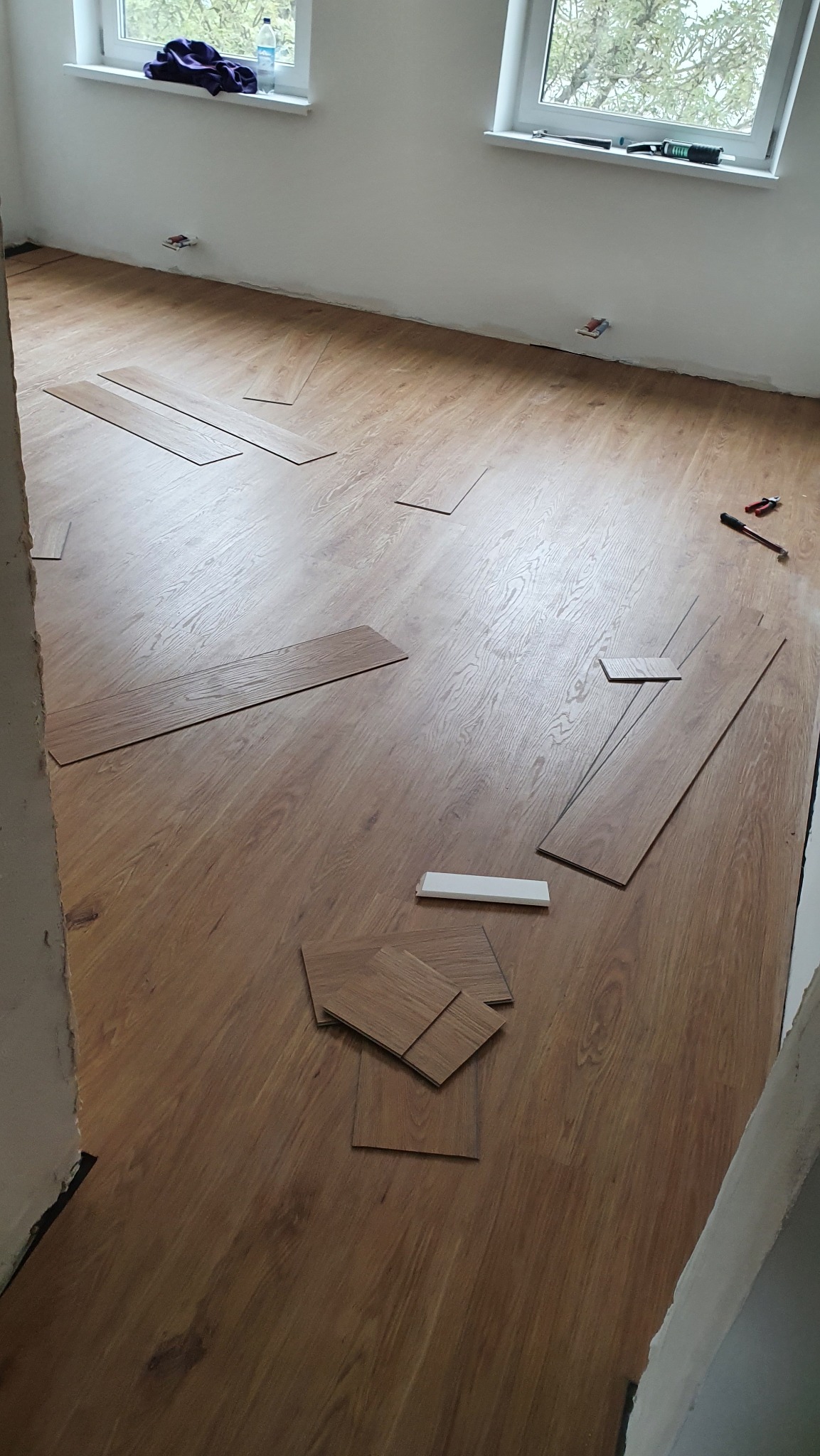 Rozpoczęte układanie paneli podłogowych w jasnym pokoju. Widoczne narzędzia i resztki paneli przy ścianie. Dwa okna w tle. Wnętrze w trakcie wykończenia.