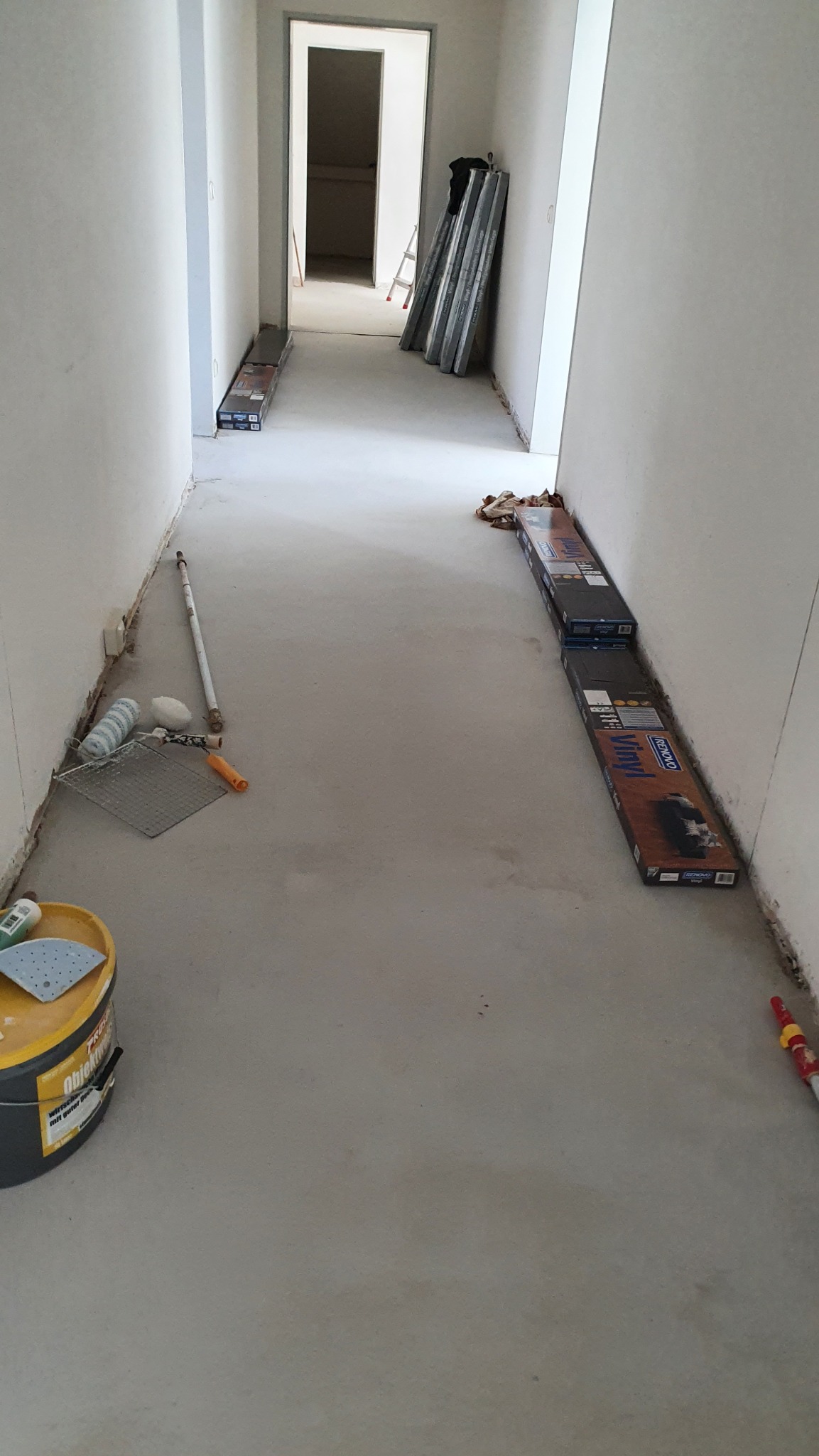 Przygotowanie do prac w korytarzu w Głogowie: puszka z farbą, wałki malarskie i panele podłogowe ułożone wzdłuż ścian. Widoczny surowy beton i drzwi w tle.