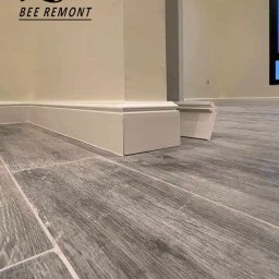 BEE REMONT ANIS DJEBAILI - Szare panele podłogowe imitujące drewno z zamontowanymi białymi listwami przypodłogowymi na ścianie z logo firmy remontowej Bee Remont.