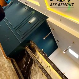 BEE REMONT ANIS DJEBAILI - Wykończone wnętrze z podwieszanym sufitem, oświetleniem LED, marmurową ścianą akcentową, pomalowaną ścianą na ciemny zielony kolor i logo firmy remontowej 'Bee Remont' na suficie.