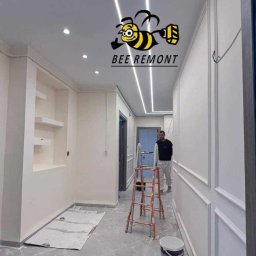 BEE REMONT ANIS DJEBAILI - Korytarz w trakcie wykańczania z dekoracyjnymi panelami ściennymi, podświetlanym sufitem i logiem firmy 'BEE REMONT' na suficie, widoczna drabina i osoba w tle.