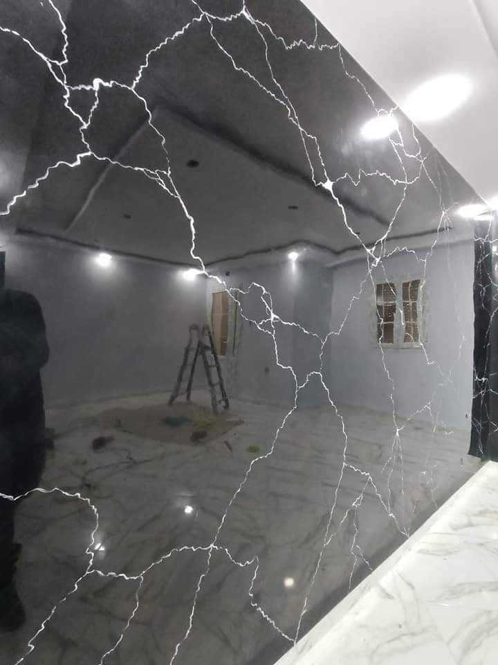 Wykończona ściana z czarnego, błyszczącego materiału imitującego marmur z białymi żyłkami, odbijająca wnętrze remontowanego pokoju z drabiną i oknem.