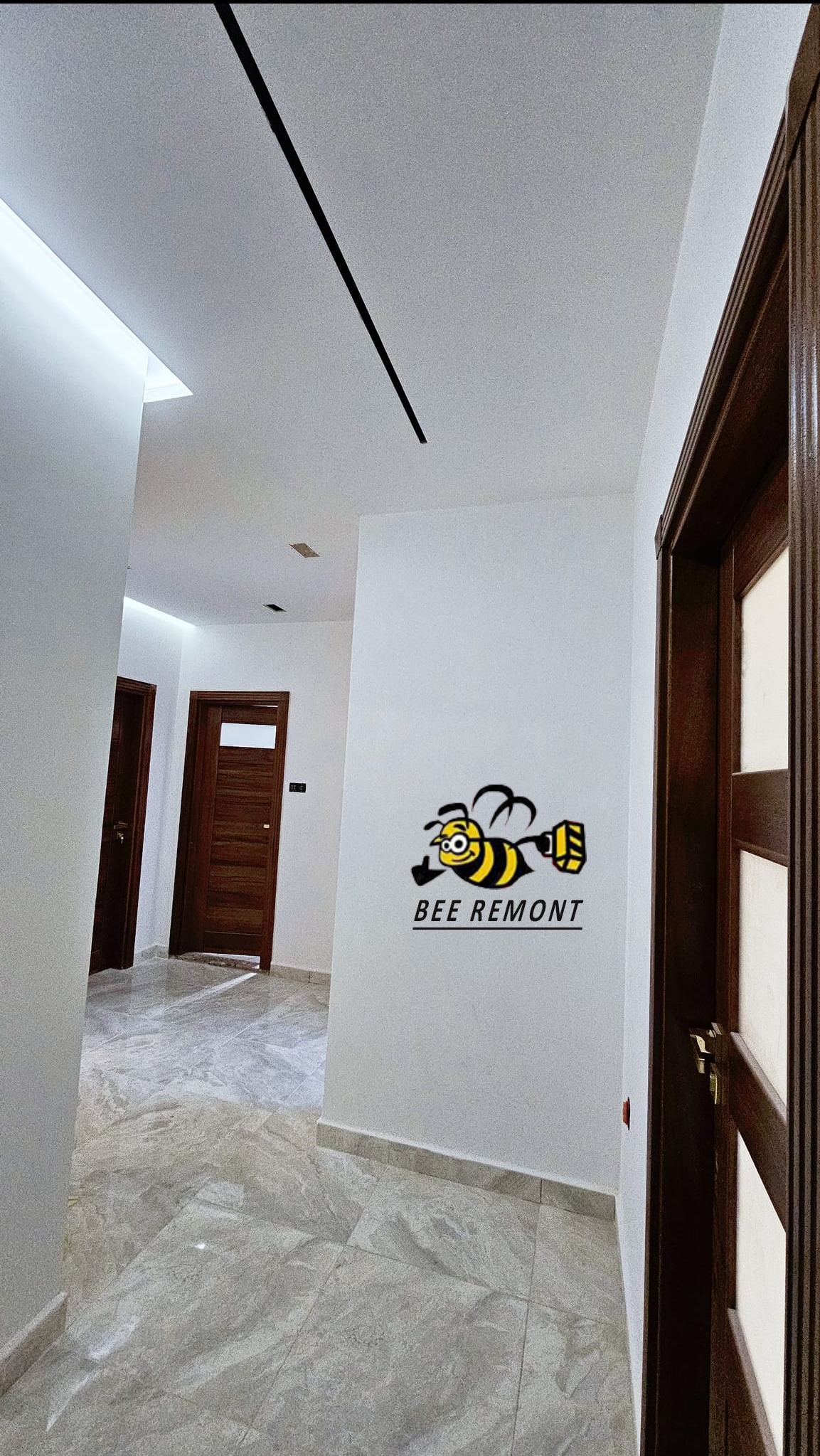 Wykończony korytarz z marmurową posadzką, białymi ścianami z logo firmy remontowej i drewnianymi drzwiami. Na suficie nowoczesne oświetlenie liniowe.