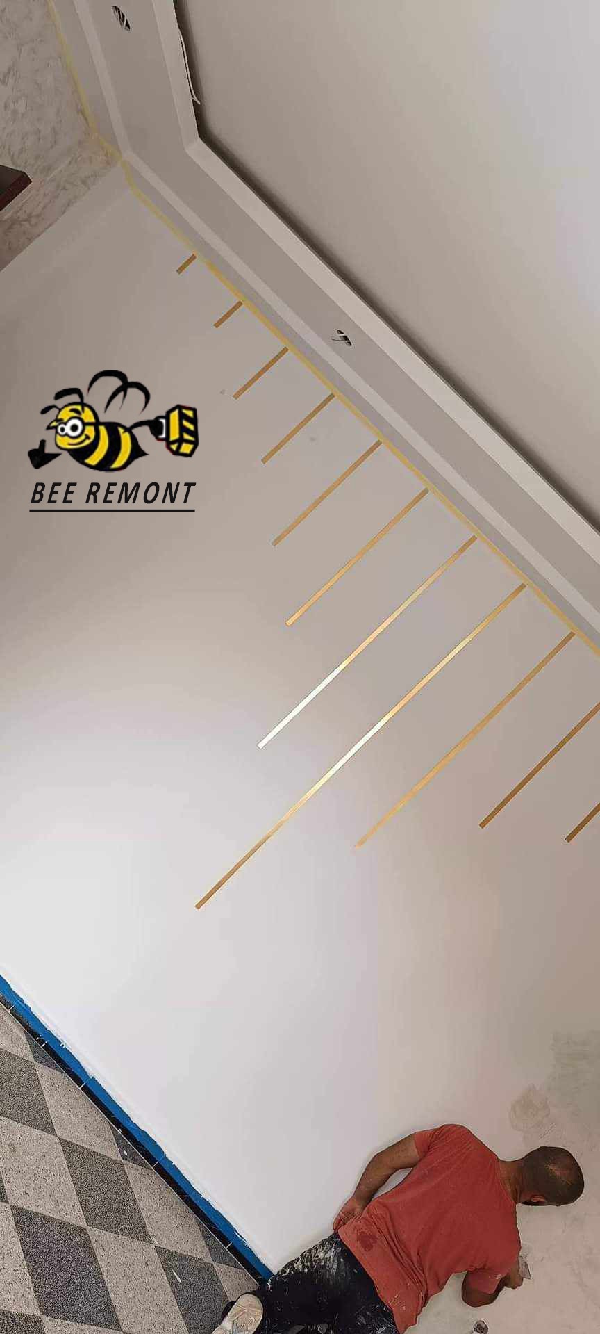 Świeżo pomalowana biała ściana z dekoracyjnymi złotymi paskami, widoczne logo firmy 'BEE REMONT' oraz fragment podłogi w szachownicę i pracownik w trakcie pracy.