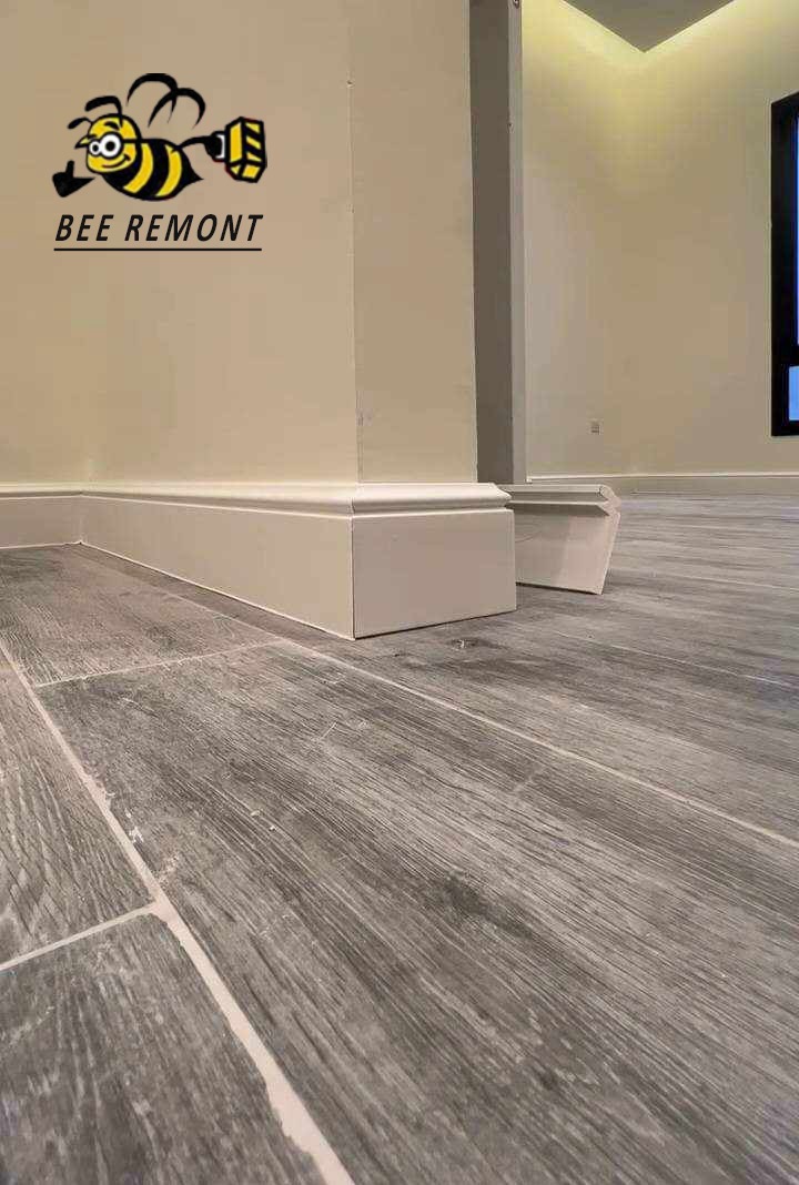 Szare panele podłogowe imitujące drewno z zamontowanymi białymi listwami przypodłogowymi na ścianie z logo firmy remontowej Bee Remont.