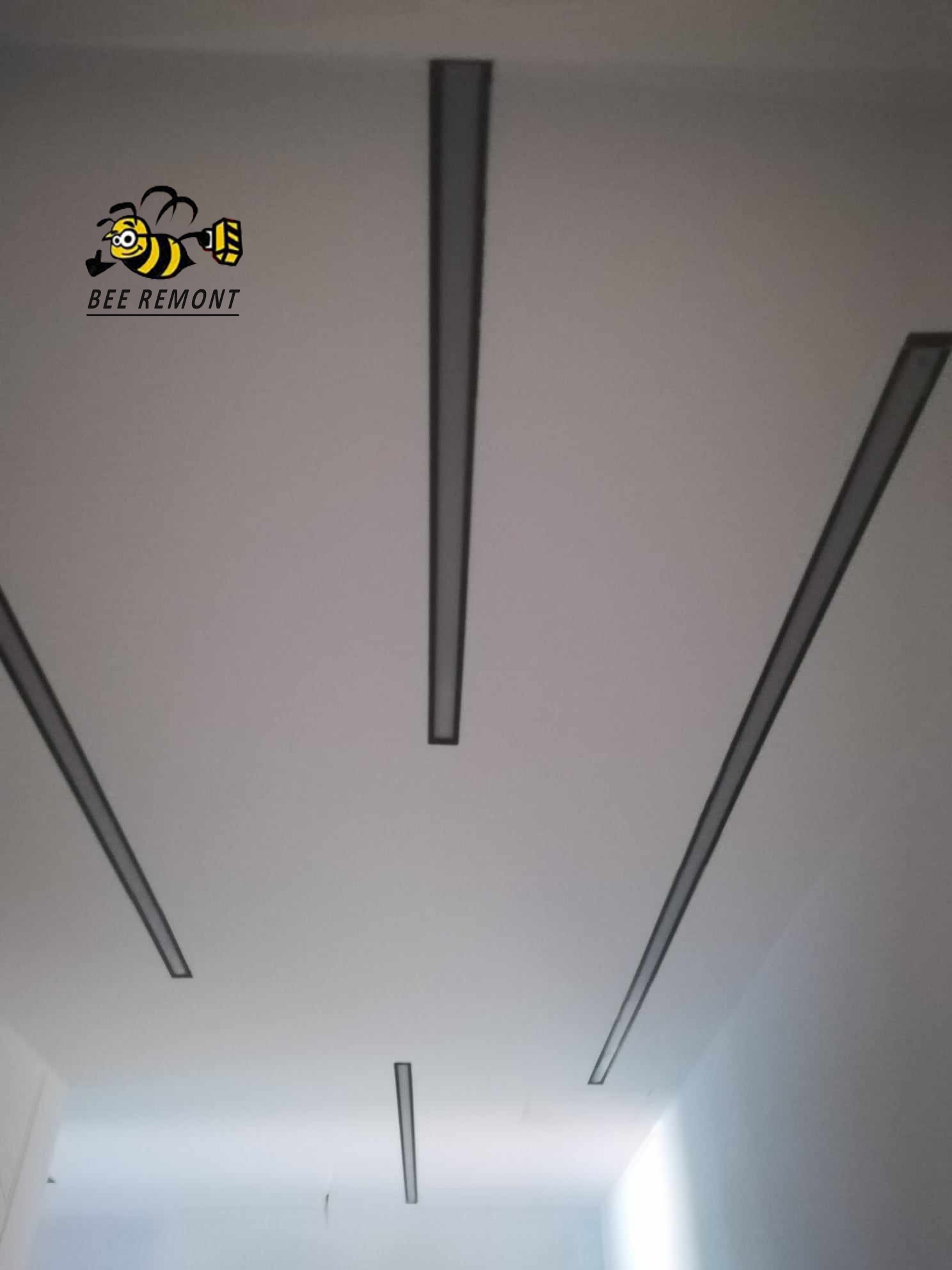 Biały sufit z zamontowanymi, podłużnymi, szarymi elementami dekoracyjnymi i widocznym logo firmy 'BEE REMONT' w lewym górnym rogu.