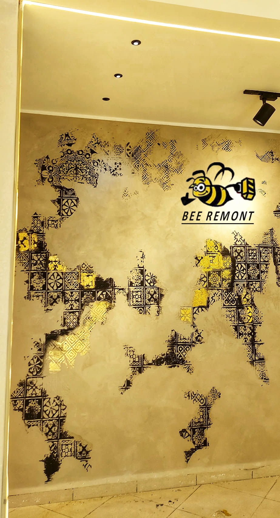 Dekoracyjna ściana z efektem postarzanego tynku, wzorami geometrycznymi w kolorze czarnym i złotym, oświetlona punktowymi lampami sufitowymi. W rogu widoczne logo firmy remontowej z wizerunkiem...