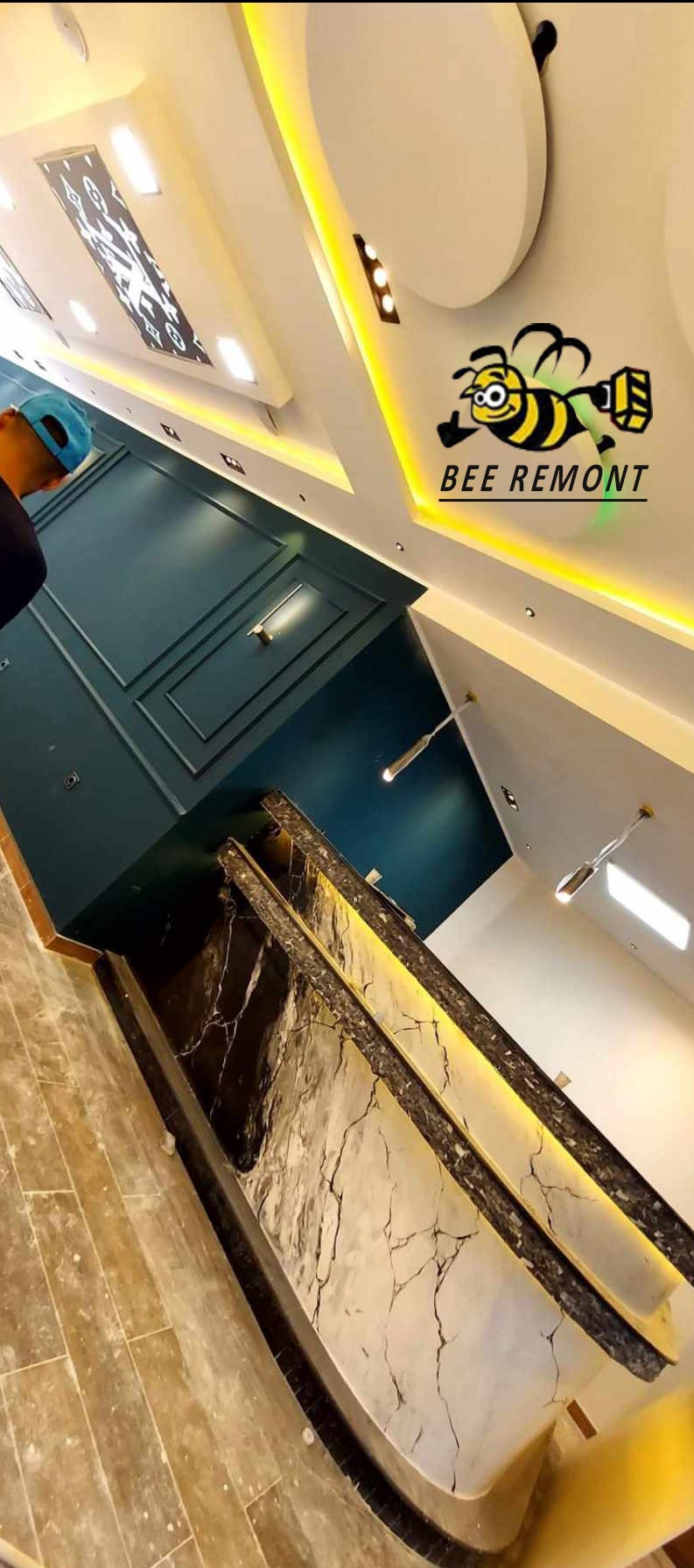 Wykończone wnętrze z podwieszanym sufitem, oświetleniem LED, marmurową ścianą akcentową, pomalowaną ścianą na ciemny zielony kolor i logo firmy remontowej 'Bee Remont' na suficie.