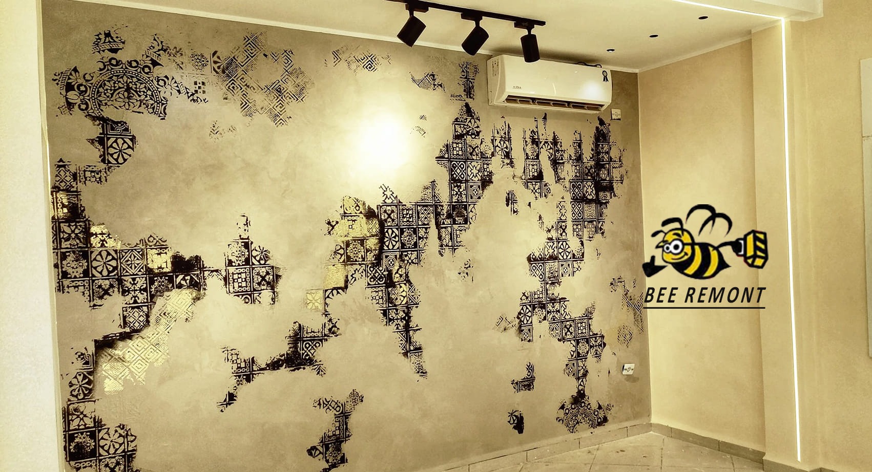 Ściana z dekoracyjną, nieregularną fakturą w odcieniach beżu i złota, pokryta czarnymi, geometrycznymi wzorami przypominającymi mozaikę, oświetlona trzema reflektorami punktowymi, z widocznym...