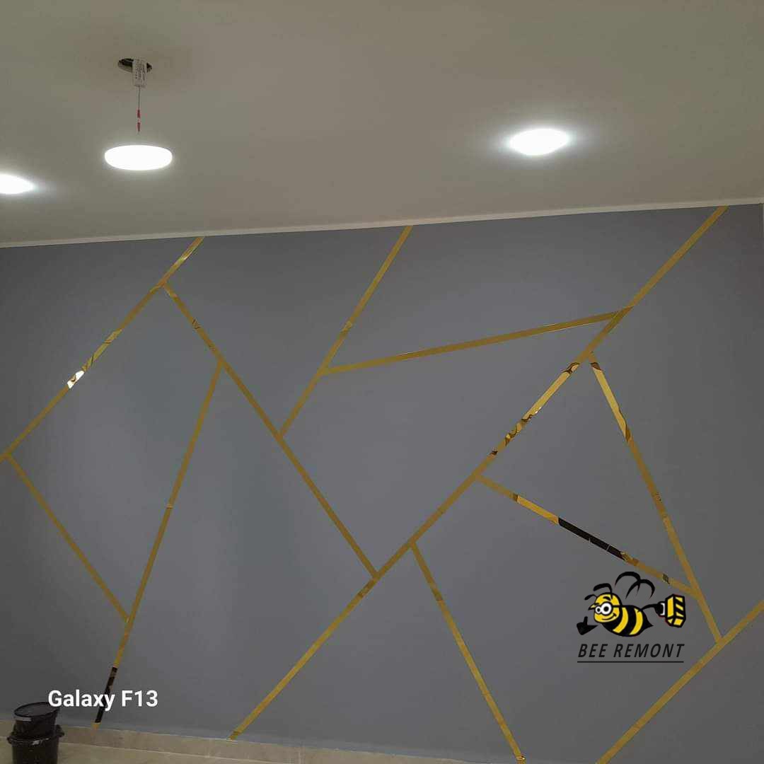 Szara ściana z geometrycznym wzorem z złotych pasków, oświetlona przez punktowe lampy sufitowe. W rogu widoczny telefon Galaxy F13 oraz logo firmy 'BEE REMONT'.