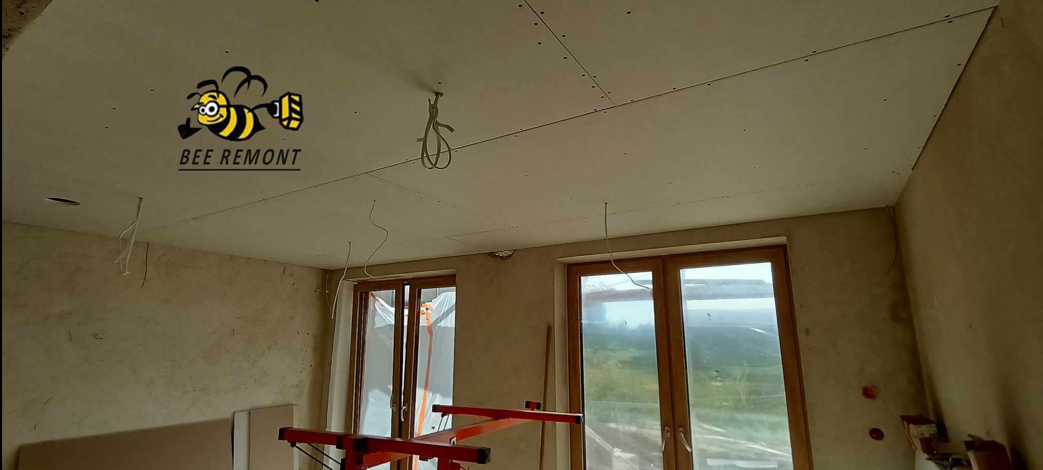 Sufit z zamontowaną płytą gipsowo-kartonową, widoczne łączenia i otwory na oświetlenie, na ścianie logo firmy remontowej w kształcie pszczoły, okna w tle.