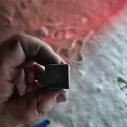 RYMER SPÓŁKA Z OGRANICZONĄ ODPOWIEDZIALNOŚCIĄ - Dłoń trzymająca szarą kostkę na tle białego, nierównego podłoża z czerwonym odblaskiem światła.