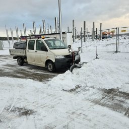 RYMER SPÓŁKA Z OGRANICZONĄ ODPOWIEDZIALNOŚCIĄ - Biała ciężarówka z pługiem odśnieżająca parking; w tle konstrukcja budynku w trakcie budowy i ogrodzenie z siatki.