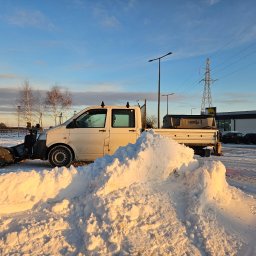 RYMER SPÓŁKA Z OGRANICZONĄ ODPOWIEDZIALNOŚCIĄ - Biały samochód dostawczy z pługiem do śniegu zaparkowany przed stertą świeżo odgarniętego śniegu w promieniach zachodzącego słońca, na tle parking z zaparkowanymi autami oraz słup wysokiego napięcia.