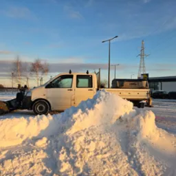 Biały samochód dostawczy z pługiem do śniegu zaparkowany przed stertą świeżo odgarniętego śniegu w promieniach zachodzącego słońca, na tle parking z zaparkowanymi autami oraz słup wysokiego napięcia.
