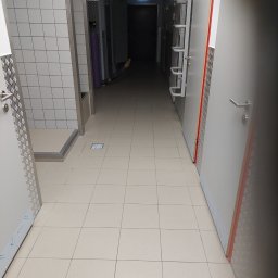 RYMER SPÓŁKA Z OGRANICZONĄ ODPOWIEDZIALNOŚCIĄ - Długi, kafelkowany korytarz z szarymi drzwiami i zielonym znakiem ewakuacyjnym w oddali, podłoga wyłożona jasnymi płytkami.