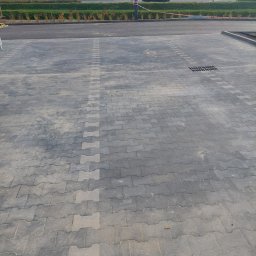 RYMER SPÓŁKA Z OGRANICZONĄ ODPOWIEDZIALNOŚCIĄ - Nowa, szara kostka brukowa ułożona na parkingu z widoczną studzienką kanalizacyjną, w tle pas zieleni i jezdnia z samochodami.