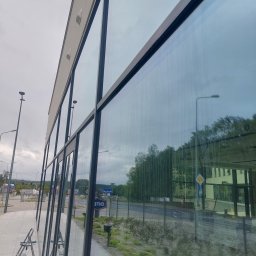 RYMER SPÓŁKA Z OGRANICZONĄ ODPOWIEDZIALNOŚCIĄ - Mycie dużych okien budynku biurowego z zewnątrz, widoczne drabiny i butelka ze środkiem czyszczącym, ślady wody na szybach.