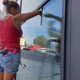 RYMER SPÓŁKA Z OGRANICZONĄ ODPOWIEDZIALNOŚCIĄ - Kobieta w czerwonym topie czyści dużą szybę za pomocą ściągaczki, widoczne smugi wody i piany, w tle czerwony van i budynek.
