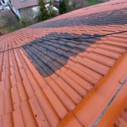 Częściowo oczyszczony dach z czerwonej dachówki, widoczna różnica między umytą a zabrudzoną powierzchnią, zarys budynków w tle.