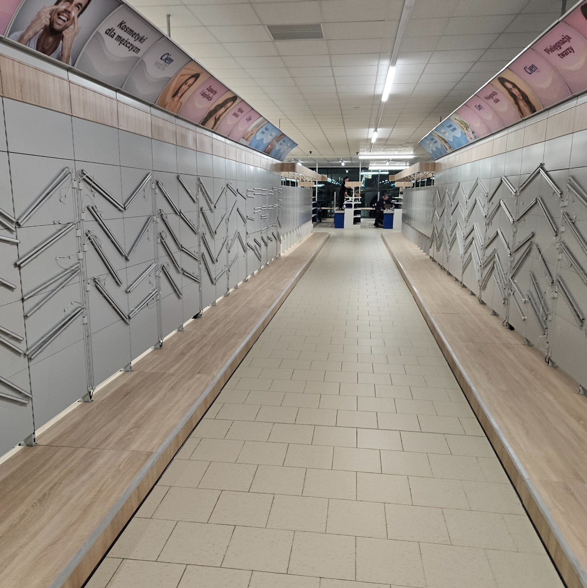 Wnętrze sklepu z pustymi półkami i reklamami kosmetyków na ścianach, widok z perspektywy podłogi wyłożonej jasnymi płytkami.