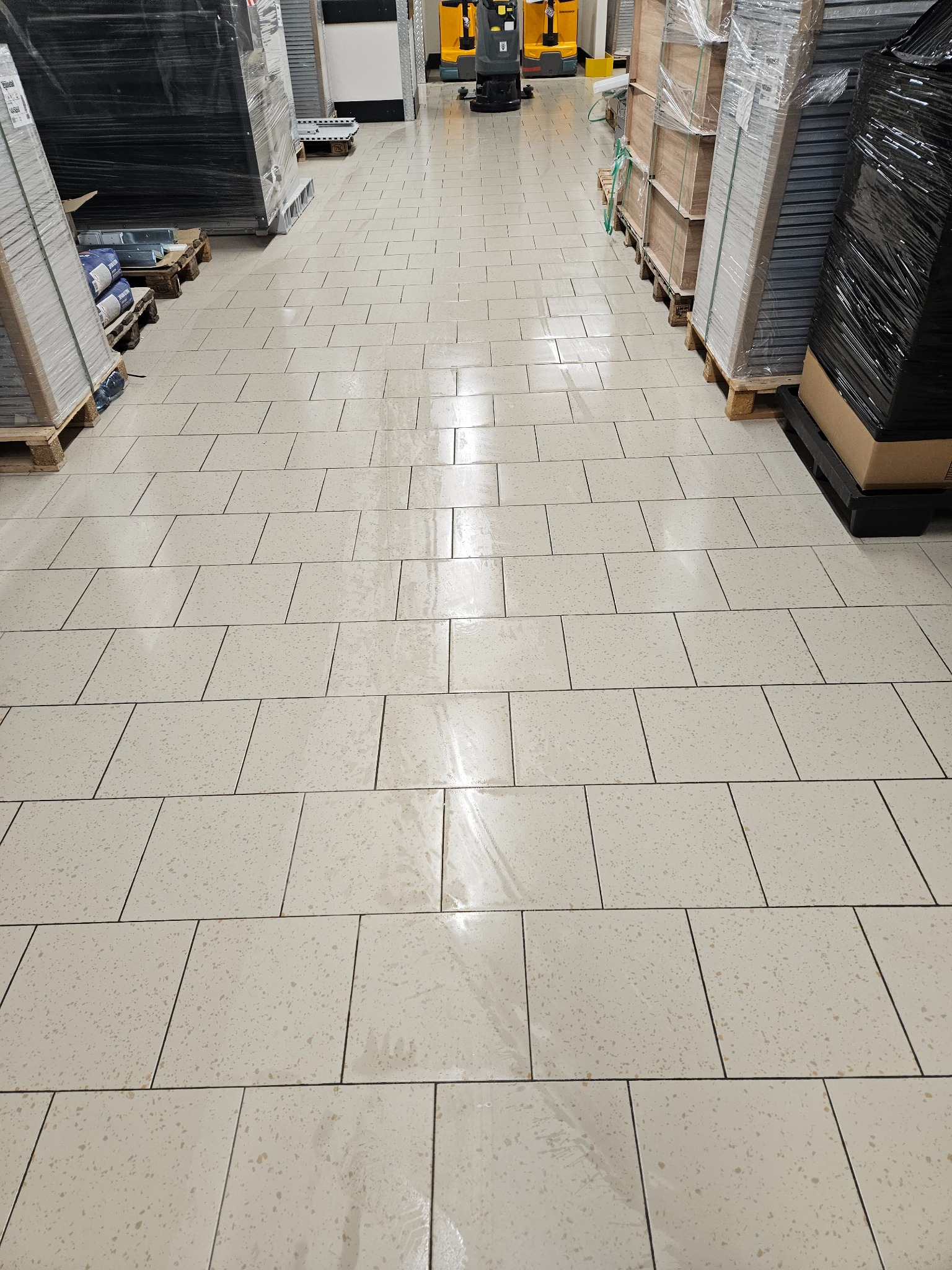 Wypolerowana, jasna podłoga z płytek w magazynie z paletami po bokach i maszyną czyszczącą w tle.