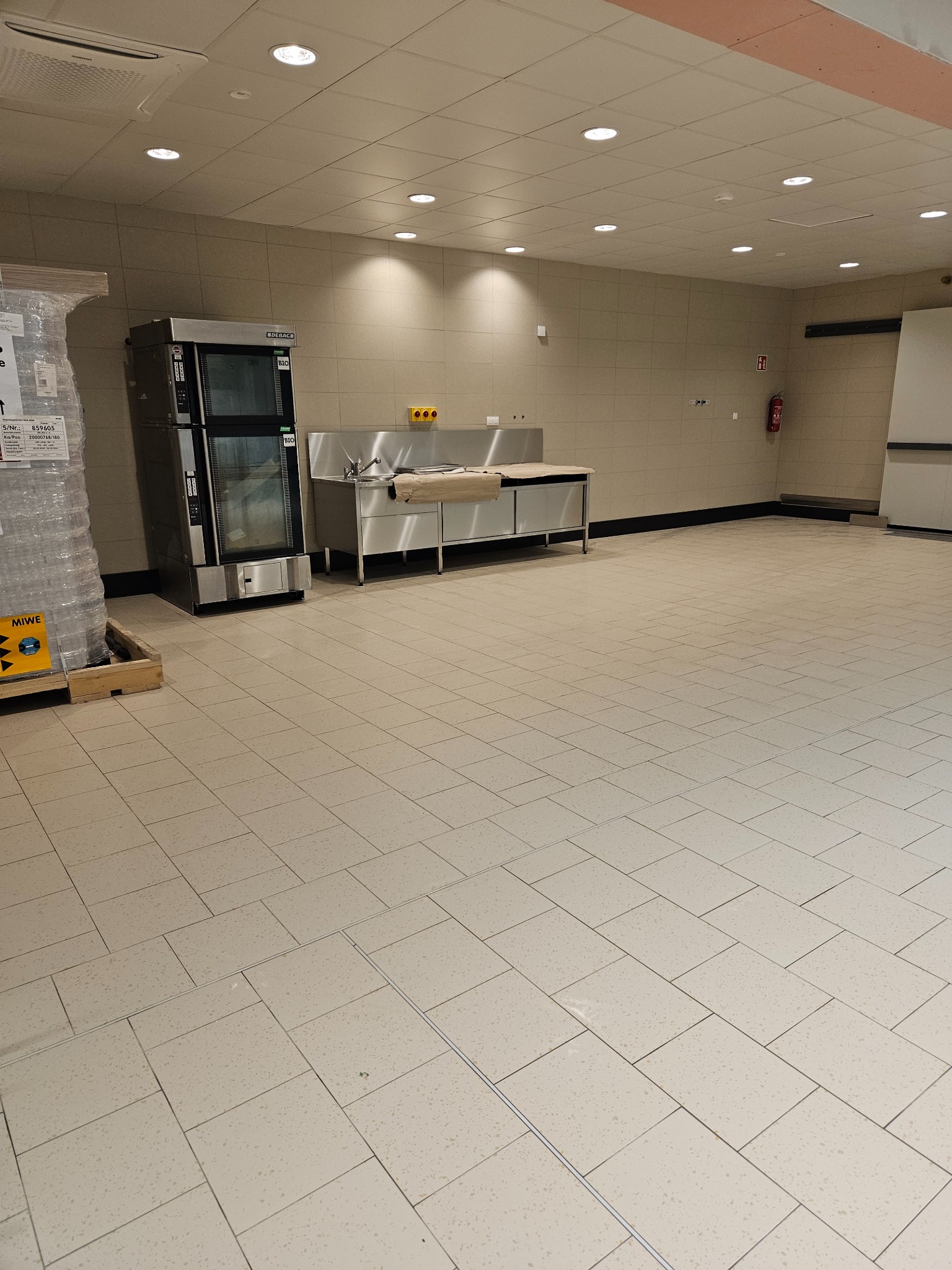 Wnętrze dużej, pustej hali z kremową terakotą na podłodze, stalową umywalką i piecem piekarniczym w tle, przygotowanej do sprzątania.