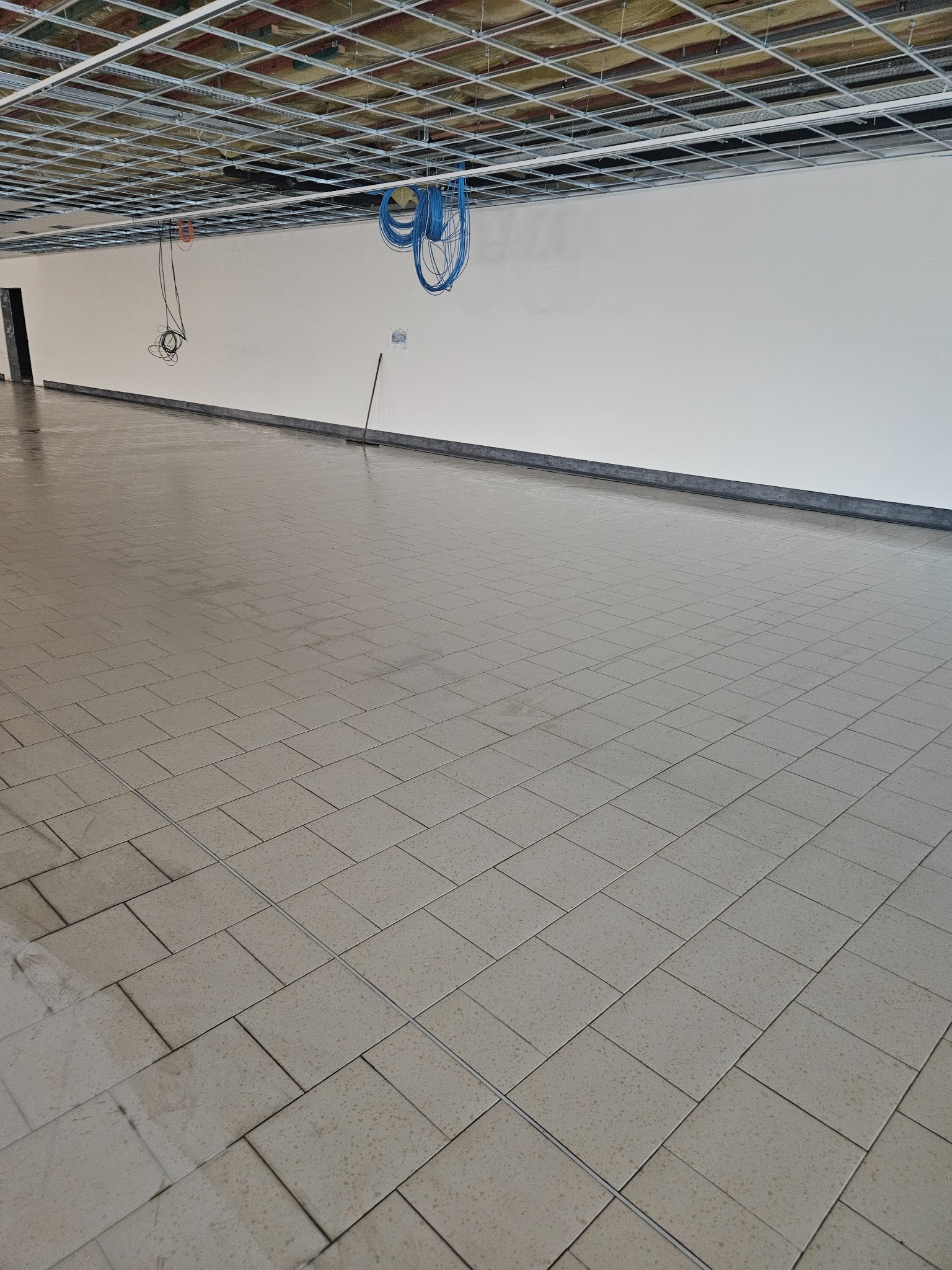 Wnętrze w trakcie remontu: surowy sufit podwieszany z widoczną konstrukcją, białe ściany, świeżo położona jasna terakota na podłodze, zwisające kable i mop oparty o ścianę.