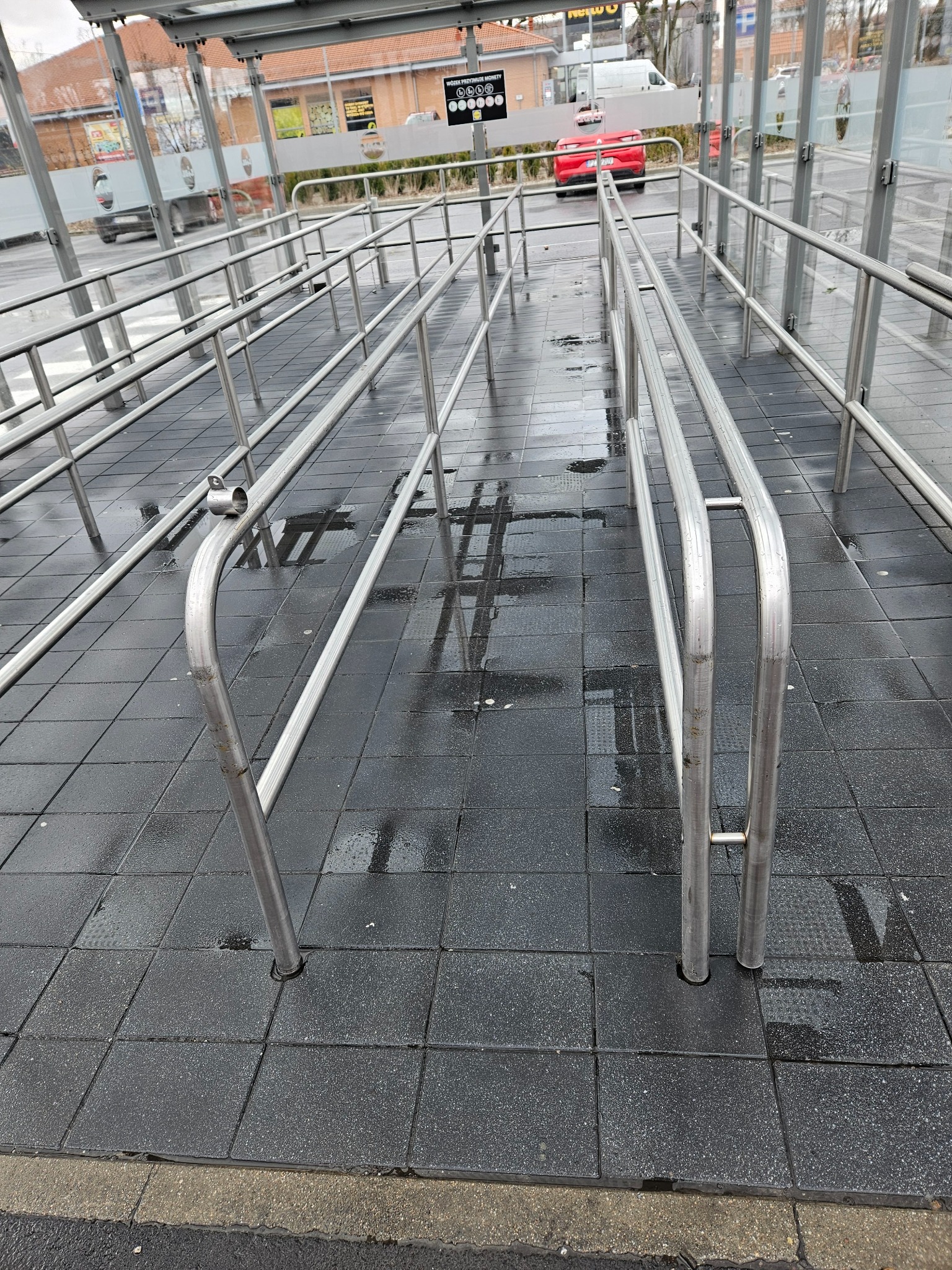 Metalowe barierki ochronne na mokrej, wyłożonej kostką placu przed budynkiem z przeszklonym przystankiem autobusowym w tle.