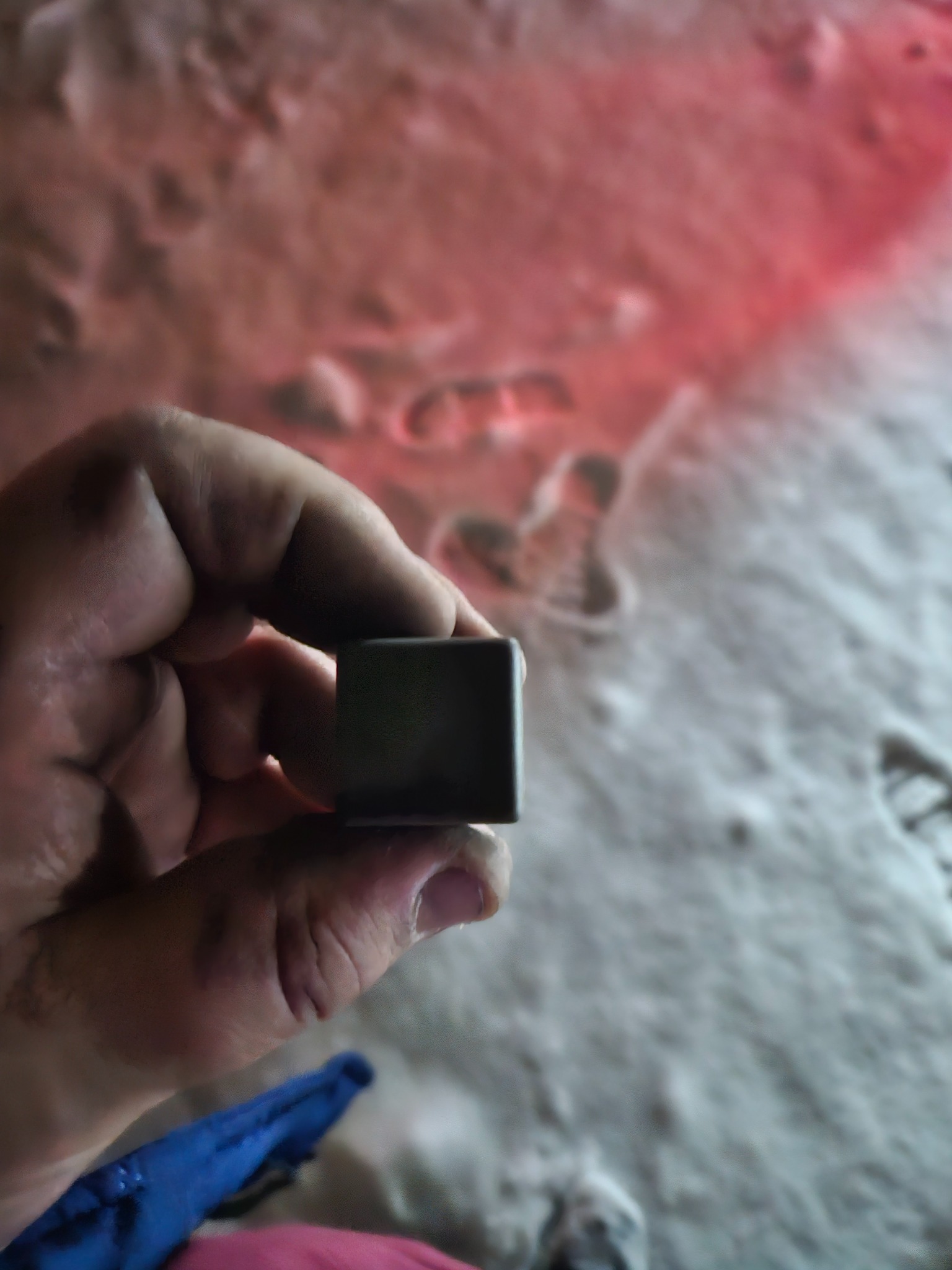 Dłoń trzymająca szarą kostkę na tle białego, nierównego podłoża z czerwonym odblaskiem światła.