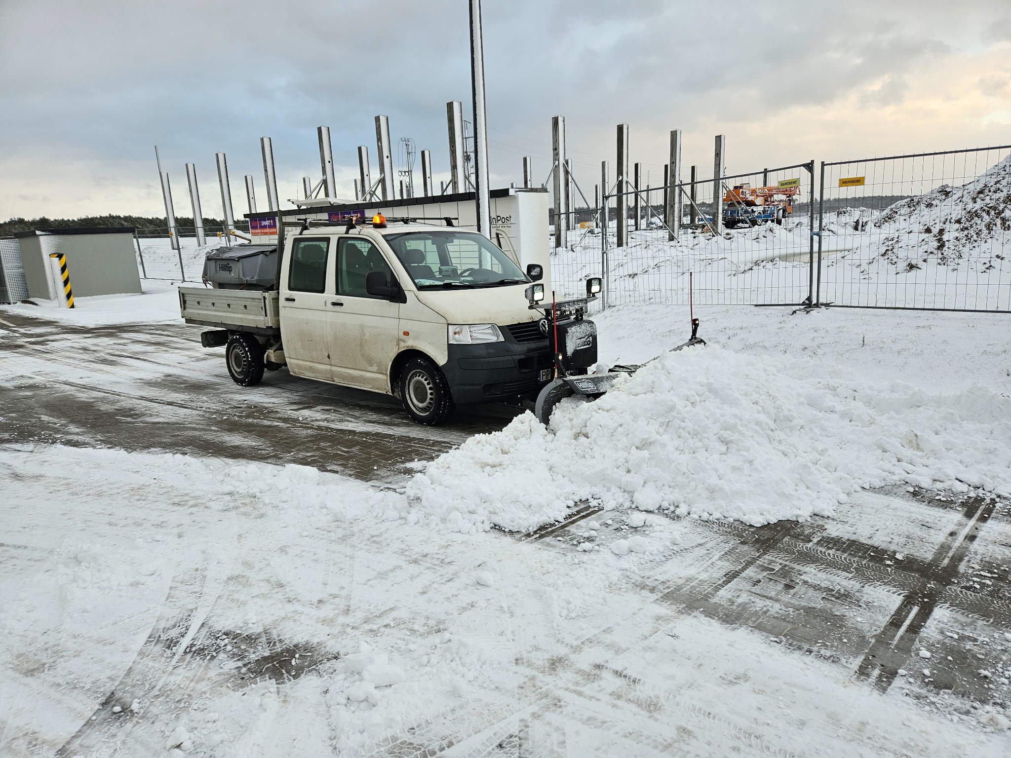 Biała ciężarówka z pługiem odśnieżająca parking; w tle konstrukcja budynku w trakcie budowy i ogrodzenie z siatki.