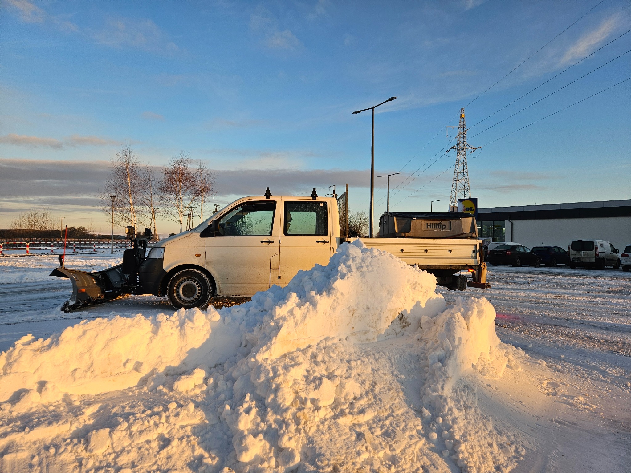 Biały samochód dostawczy z pługiem do śniegu zaparkowany przed stertą świeżo odgarniętego śniegu w promieniach zachodzącego słońca, na tle parking z zaparkowanymi autami oraz słup wysokiego napięcia.