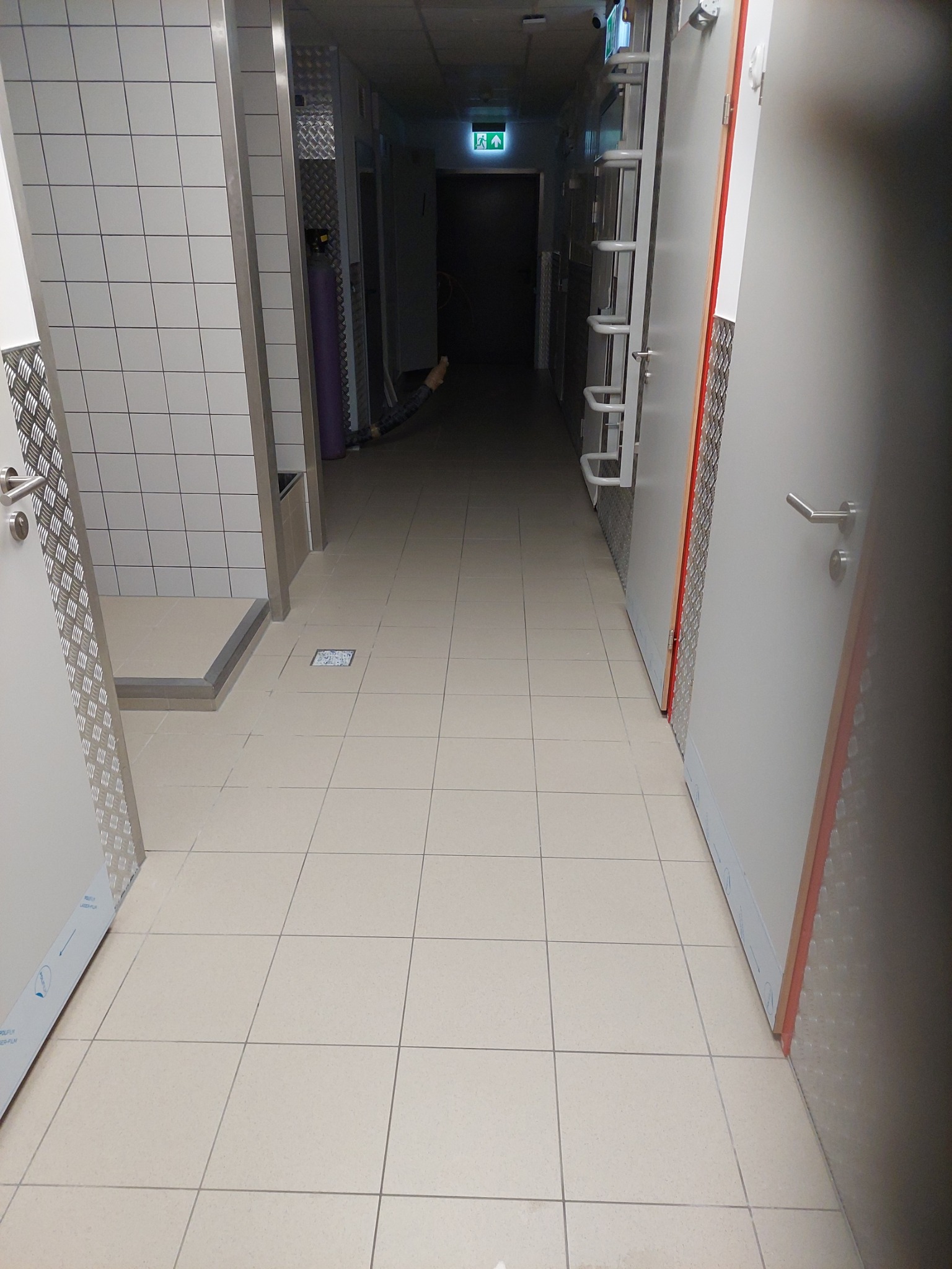 Długi, kafelkowany korytarz z szarymi drzwiami i zielonym znakiem ewakuacyjnym w oddali, podłoga wyłożona jasnymi płytkami.
