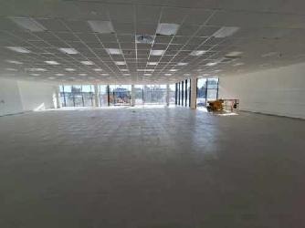 Pusta, jasna hala biurowa z betonową podłogą, widoczne okna na całej ścianie i system oświetlenia sufitowego.