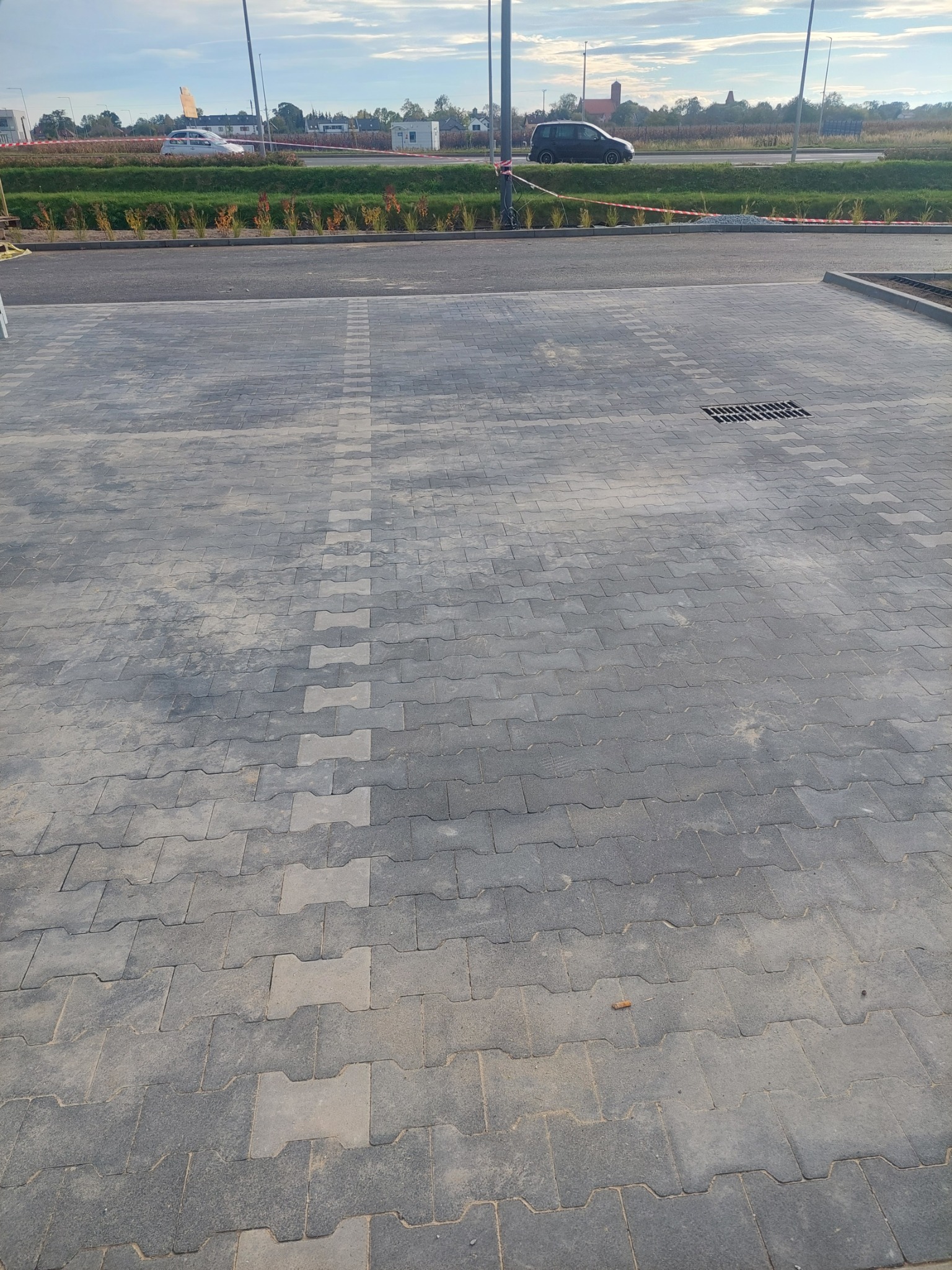 Nowa, szara kostka brukowa ułożona na parkingu z widoczną studzienką kanalizacyjną, w tle pas zieleni i jezdnia z samochodami.
