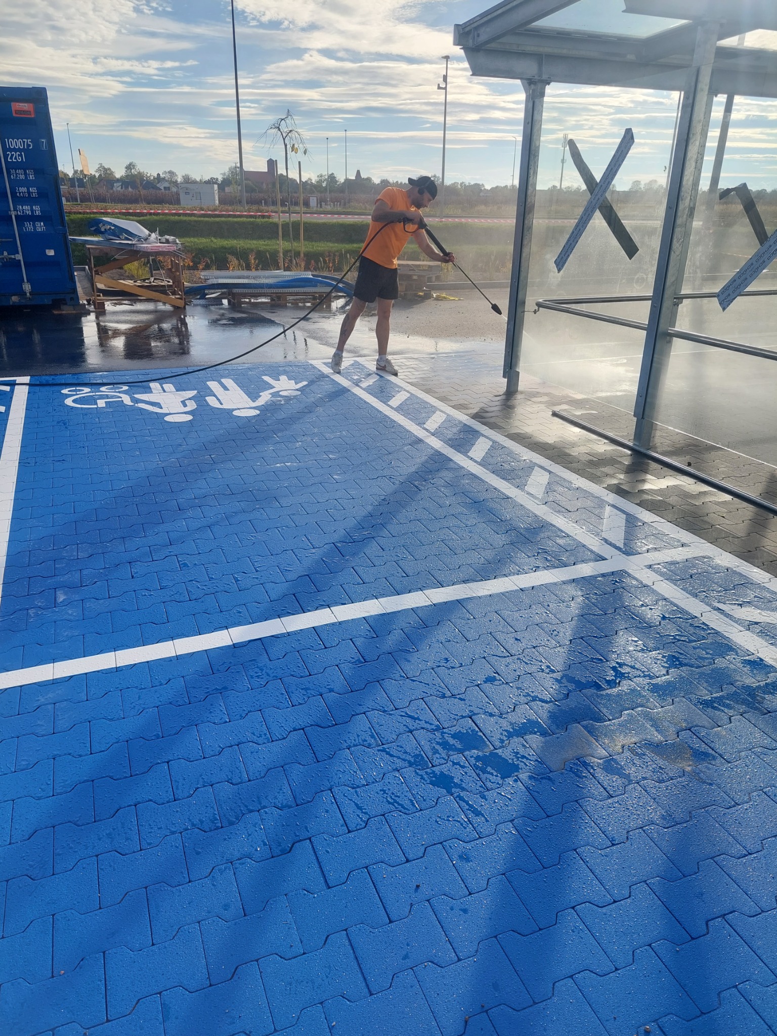 Mężczyzna w pomarańczowej koszulce czyści ciśnieniowo niebieską nawierzchnię parkingu z białymi symbolami miejsc dla rodzin z dziećmi, w tle widoczna wiata przystankowa i budynki.