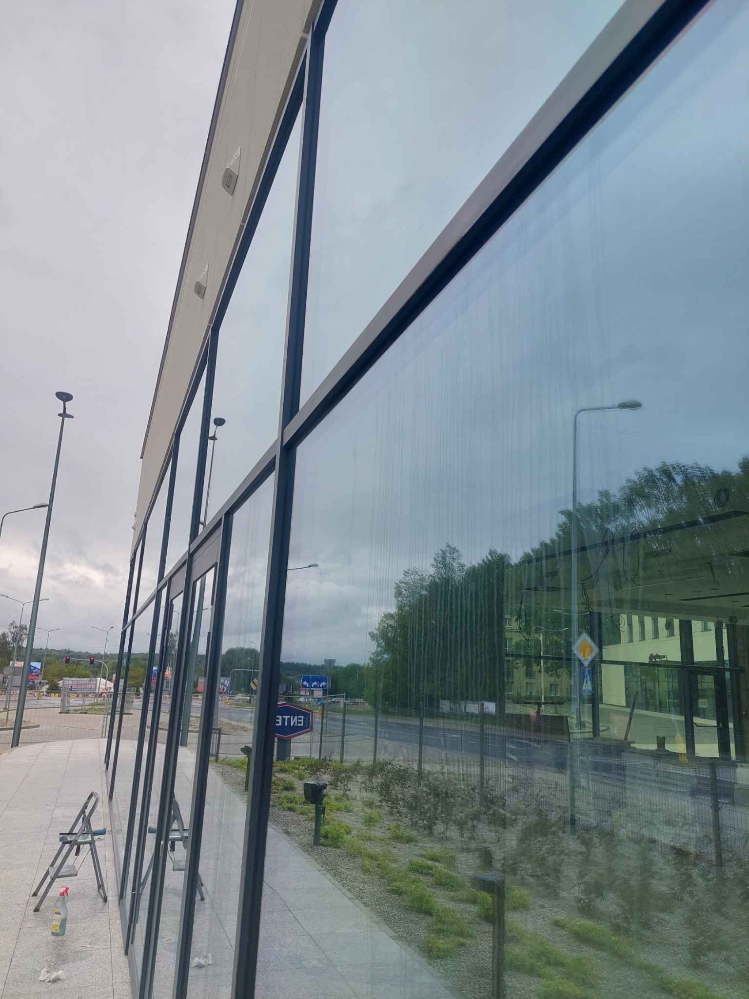 Mycie dużych okien budynku biurowego z zewnątrz, widoczne drabiny i butelka ze środkiem czyszczącym, ślady wody na szybach.